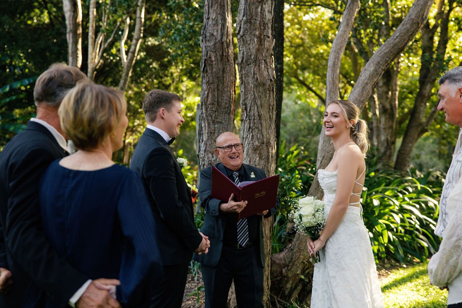 Outdoor wedding ceremony in lush garden with bride holding bouquet, groom, and officiant reading vows beneath tall trees.