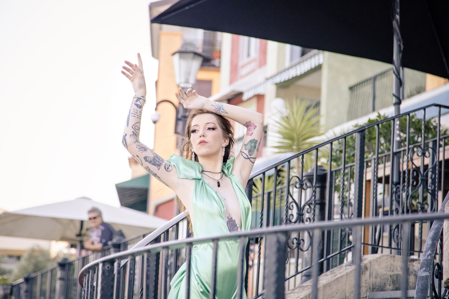 A person in a mint green outfit stands on a balcony with raised arms against an urban backdrop.