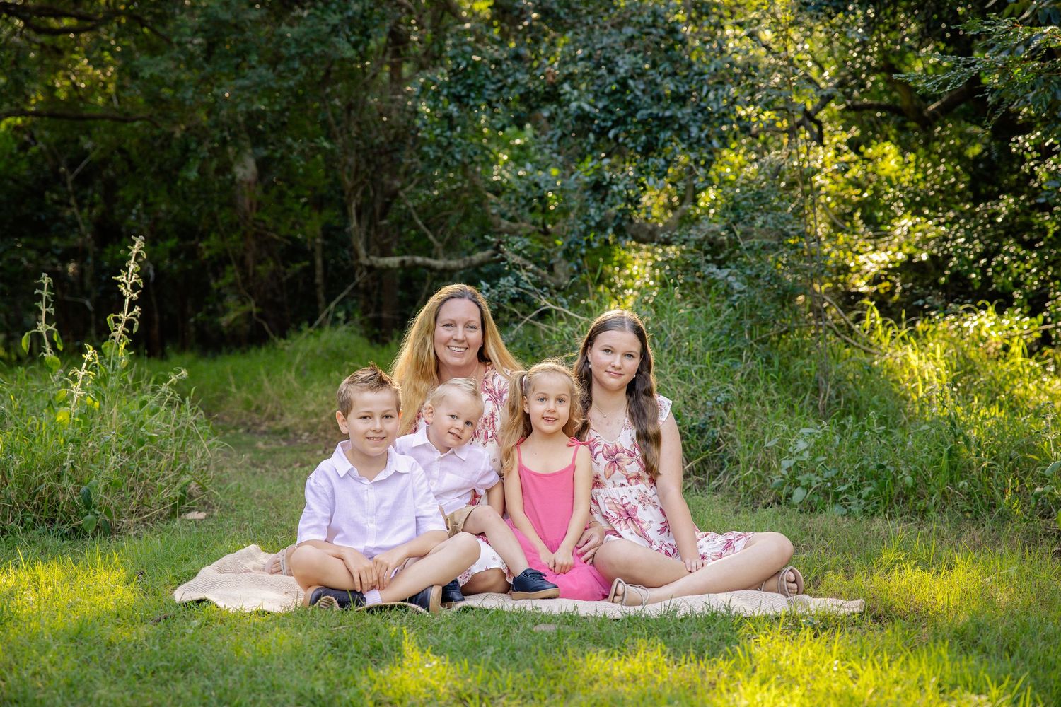 Children laying on a white blanket in a sunny meadow surrounded by green trees and natural light.