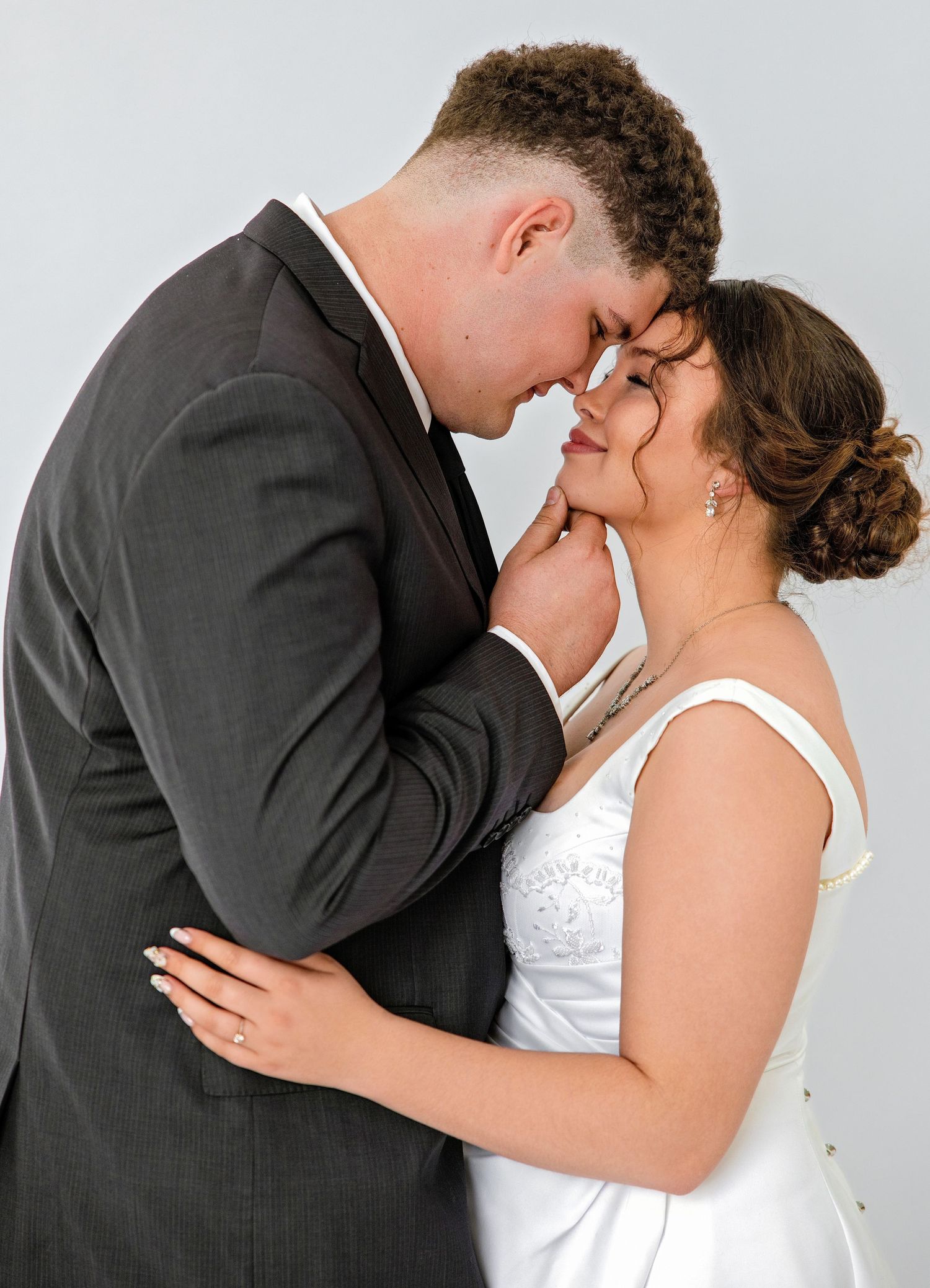 A bride and groom share a romantic forehead touch, dressed in a white gown and gray suit, in a professional wedding portrait.