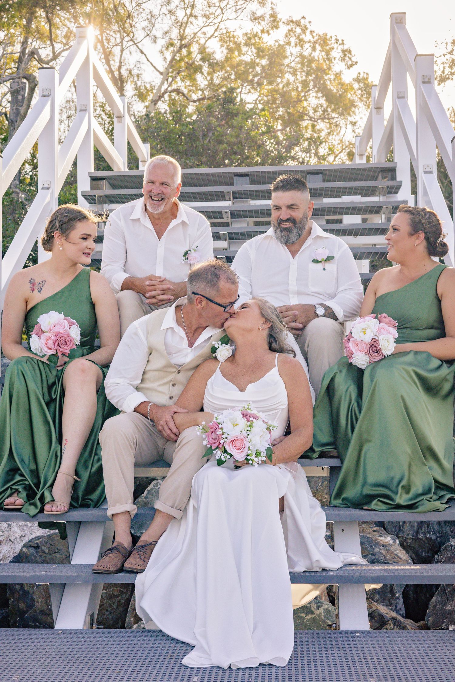 Wedding party in green bridesmaid dresses and beige suits sit together on white wooden steps during sunset.