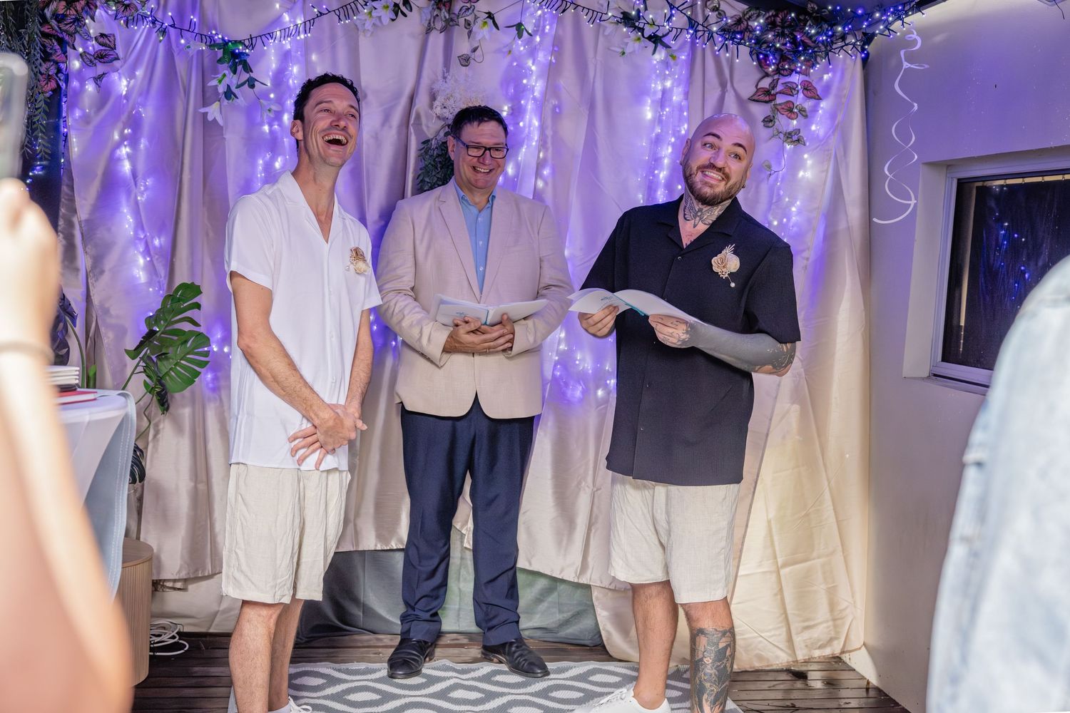 Three people share a lighthearted moment during a wedding ceremony with purple lighting and white decor.