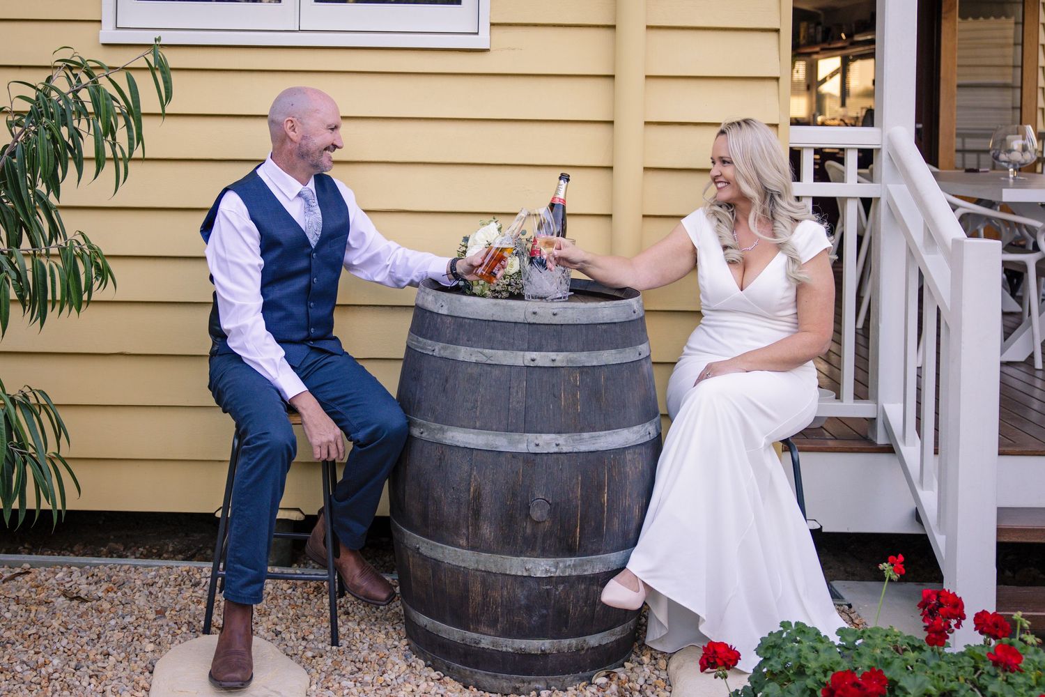 A couple in wedding attire sits on opposite sides of a large wine barrel on a porch with red flowers.