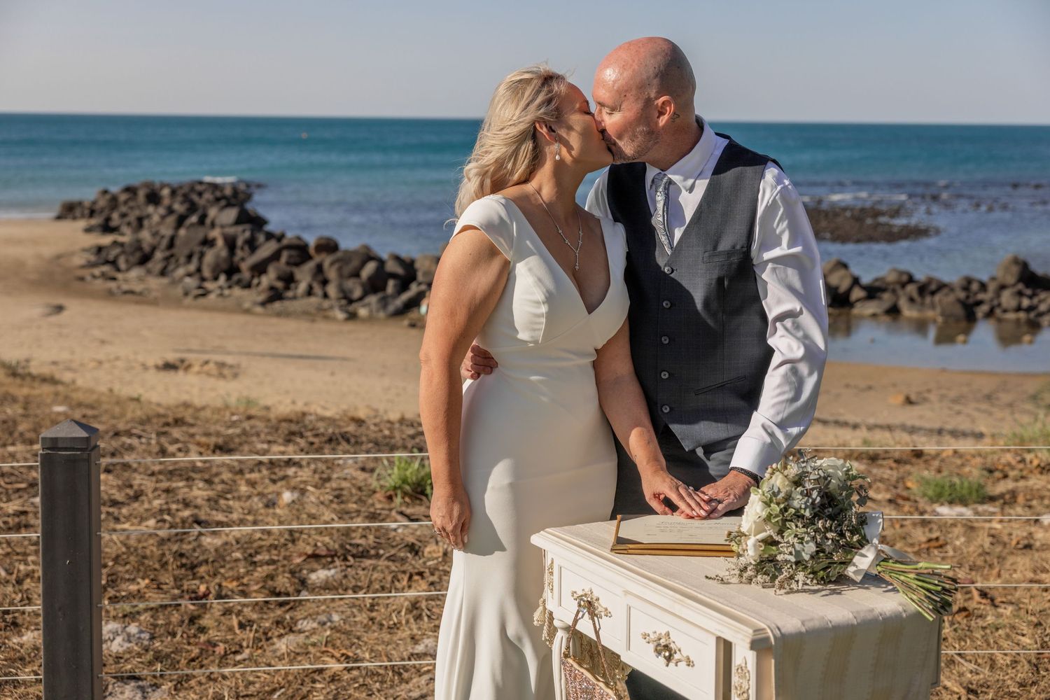 A couple shares a romantic moment on a beach during their wedding ceremony beside a vintage table with flowers.