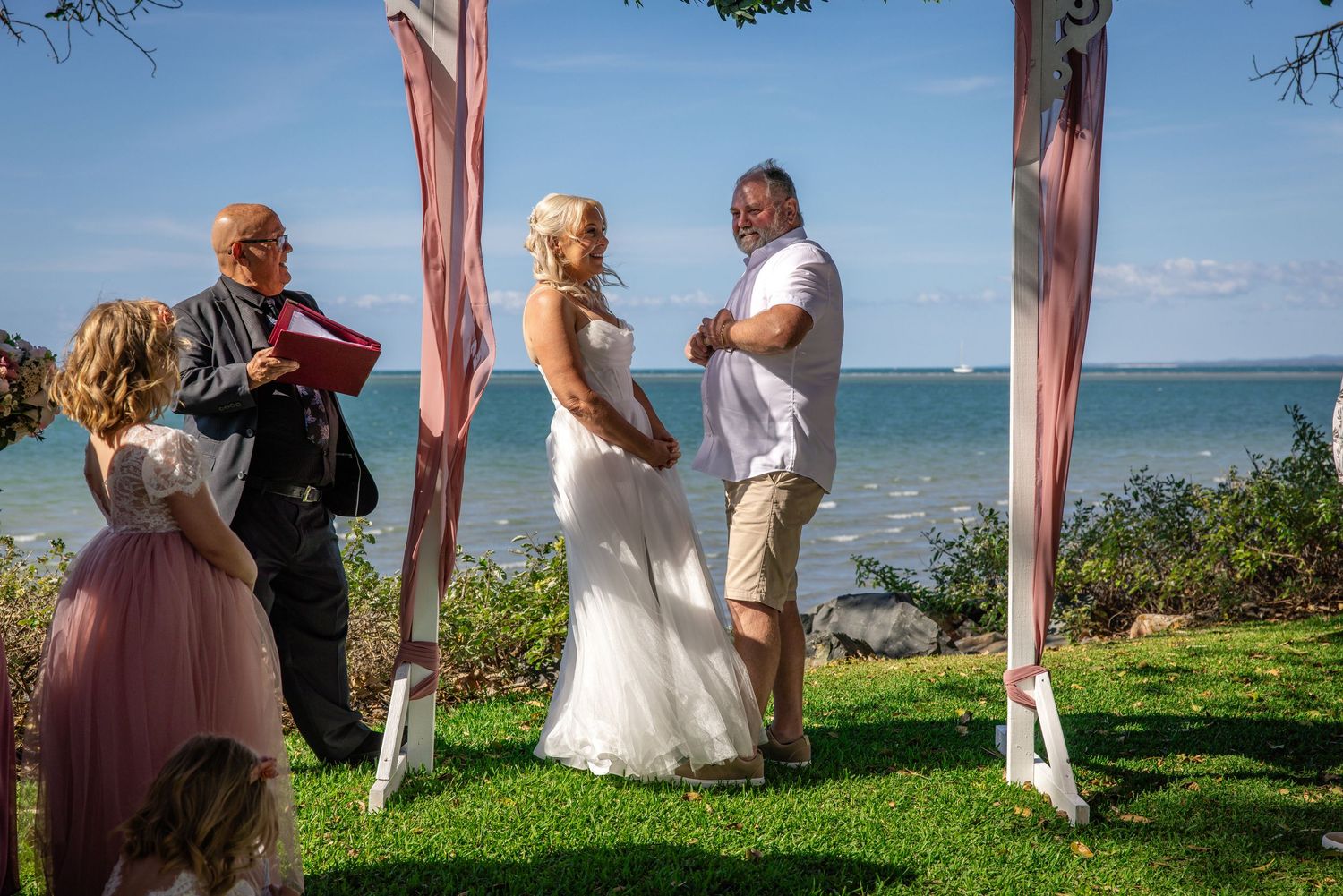 A wedding ceremony takes place outdoors with pink drapes and the ocean as a backdrop on a sunny day.