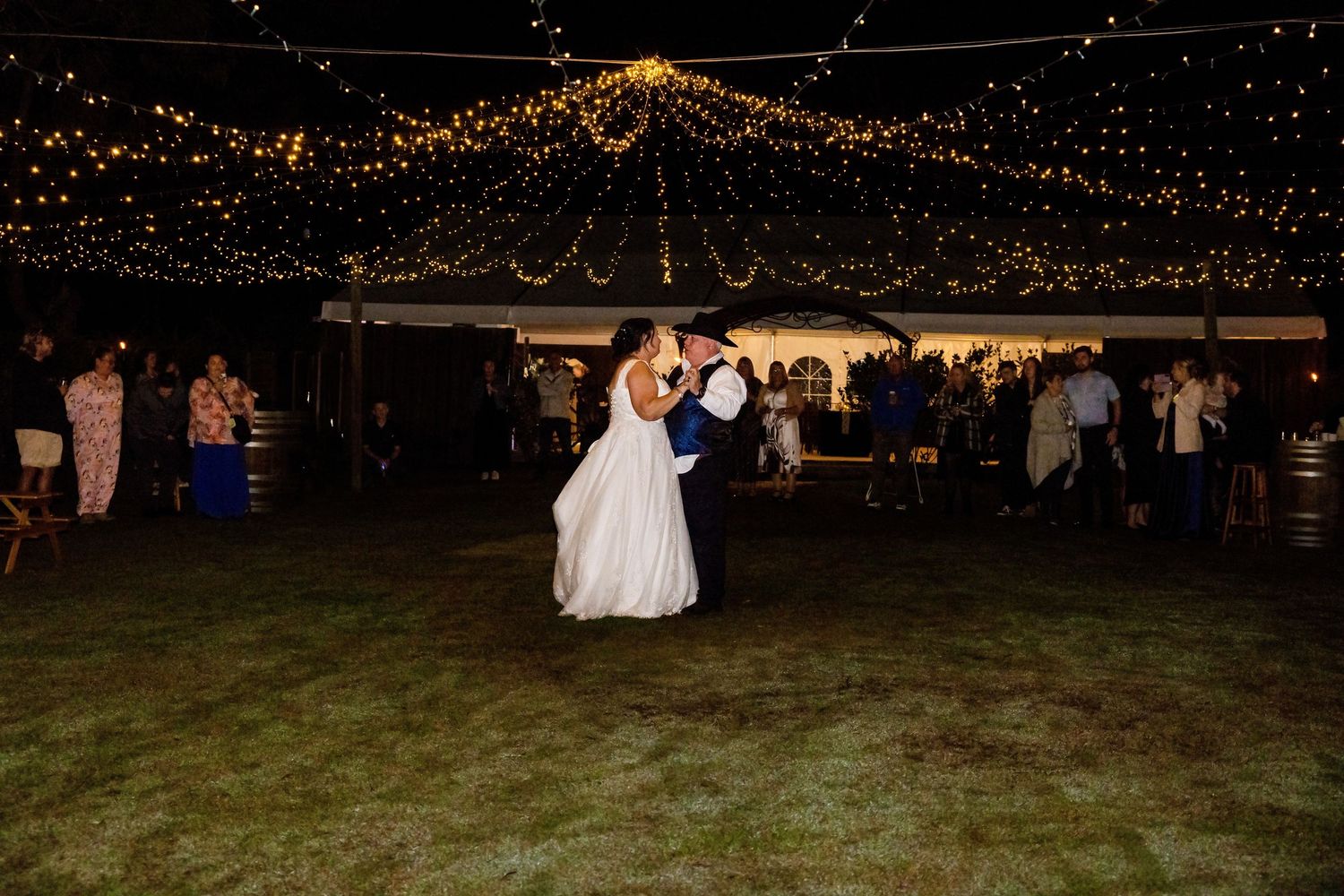 Bride and groom share first dance under twinkling fairy lights at outdoor wedding reception on lush green lawn.