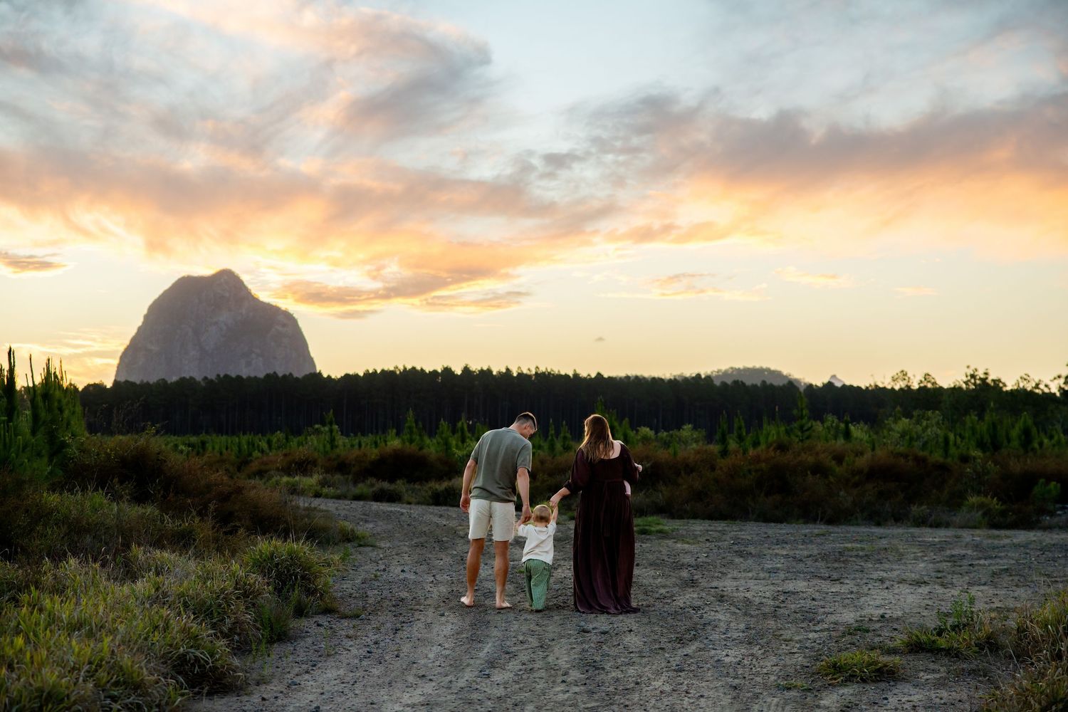 Family of three watching sunset over rocky mountain landscape on a dirt path surrounded by lush greenery.