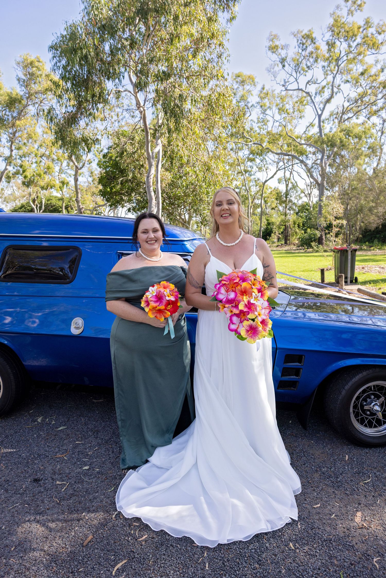 A wedding photo showing a classic blue vintage car alongside two people holding vibrant coral and pink bouquets.