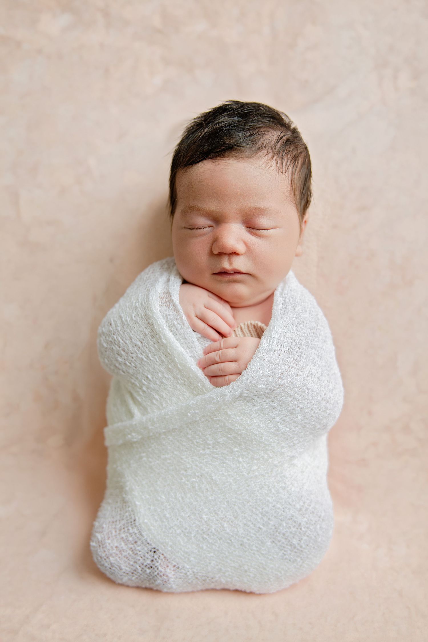 Sleeping newborn baby swaddled in white wrap on beige background, peaceful infant portrait photography.