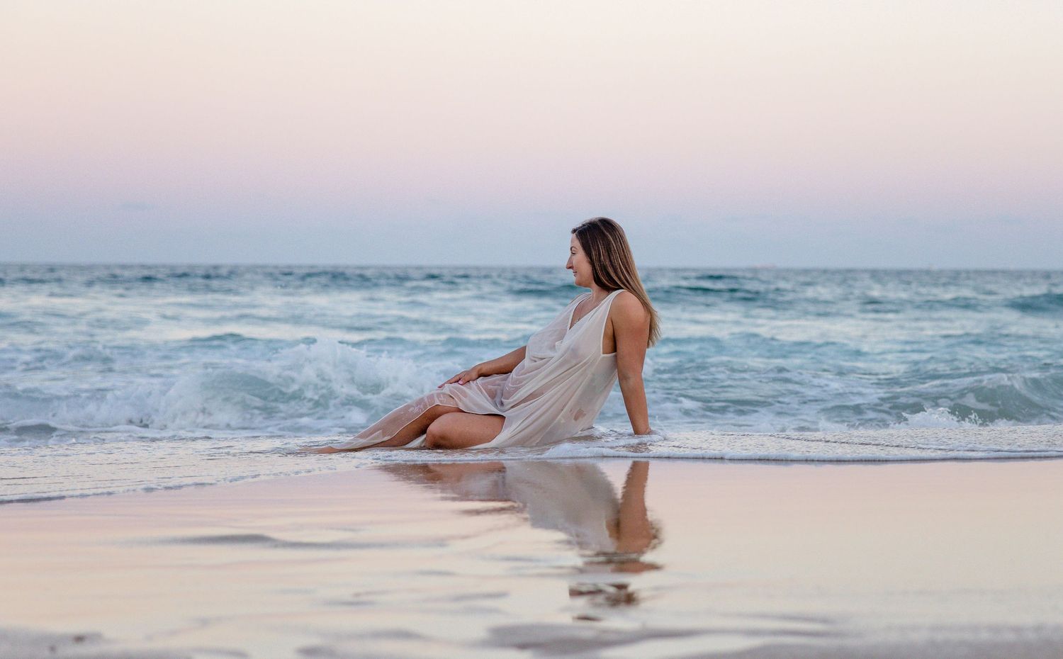 A person sits peacefully on a sandy beach at sunset while gentle waves roll in from the ocean.