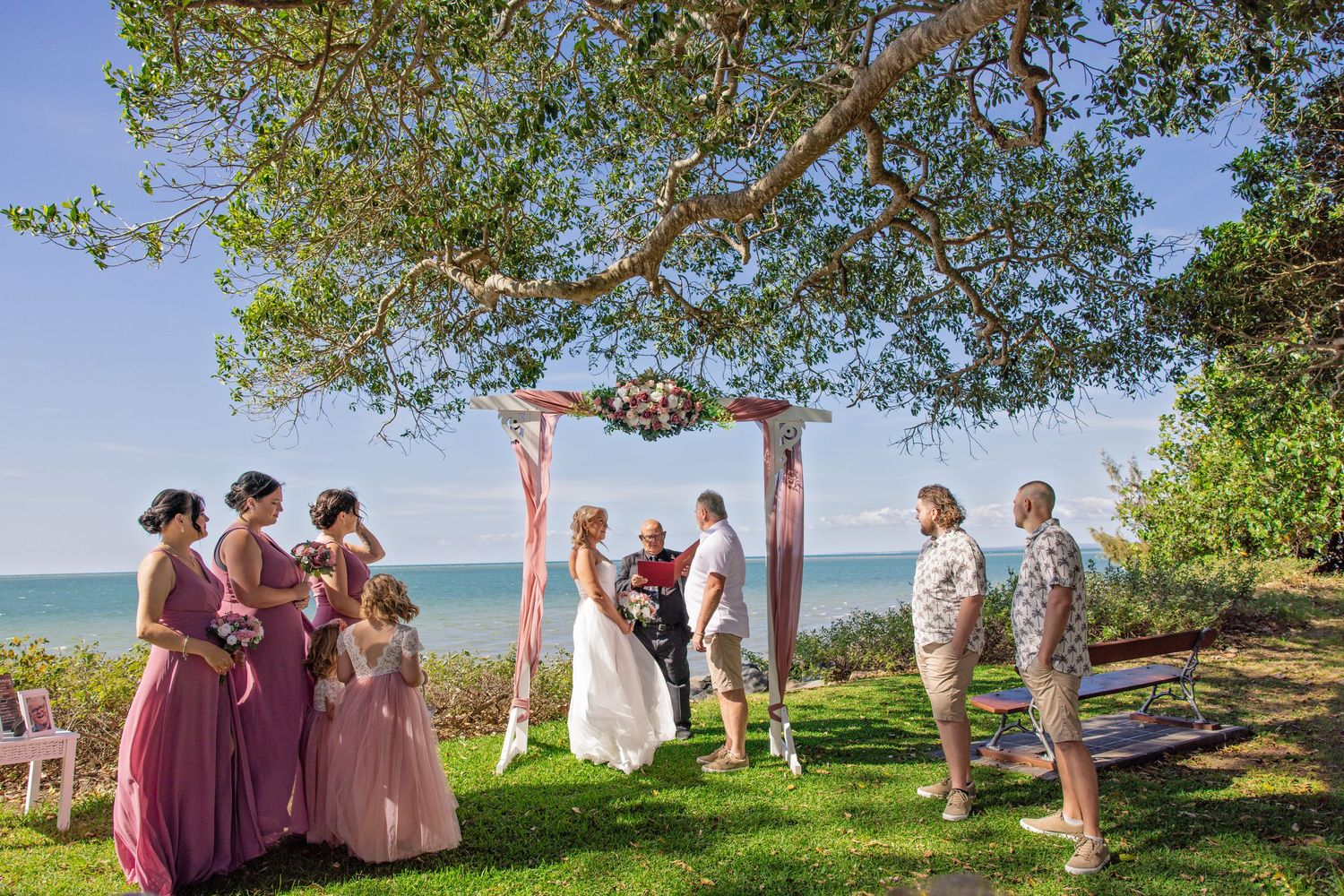 Outdoor wedding ceremony under a wooden arch with pink fabric draping, overlooking water on a sunny day.