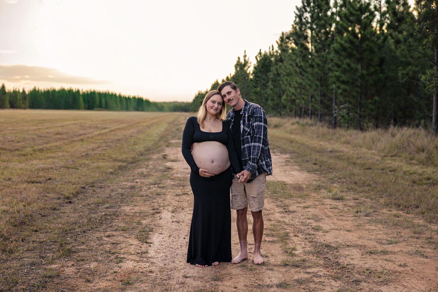 Two people pose along a winding dirt road through rolling hills and fields at sunset.