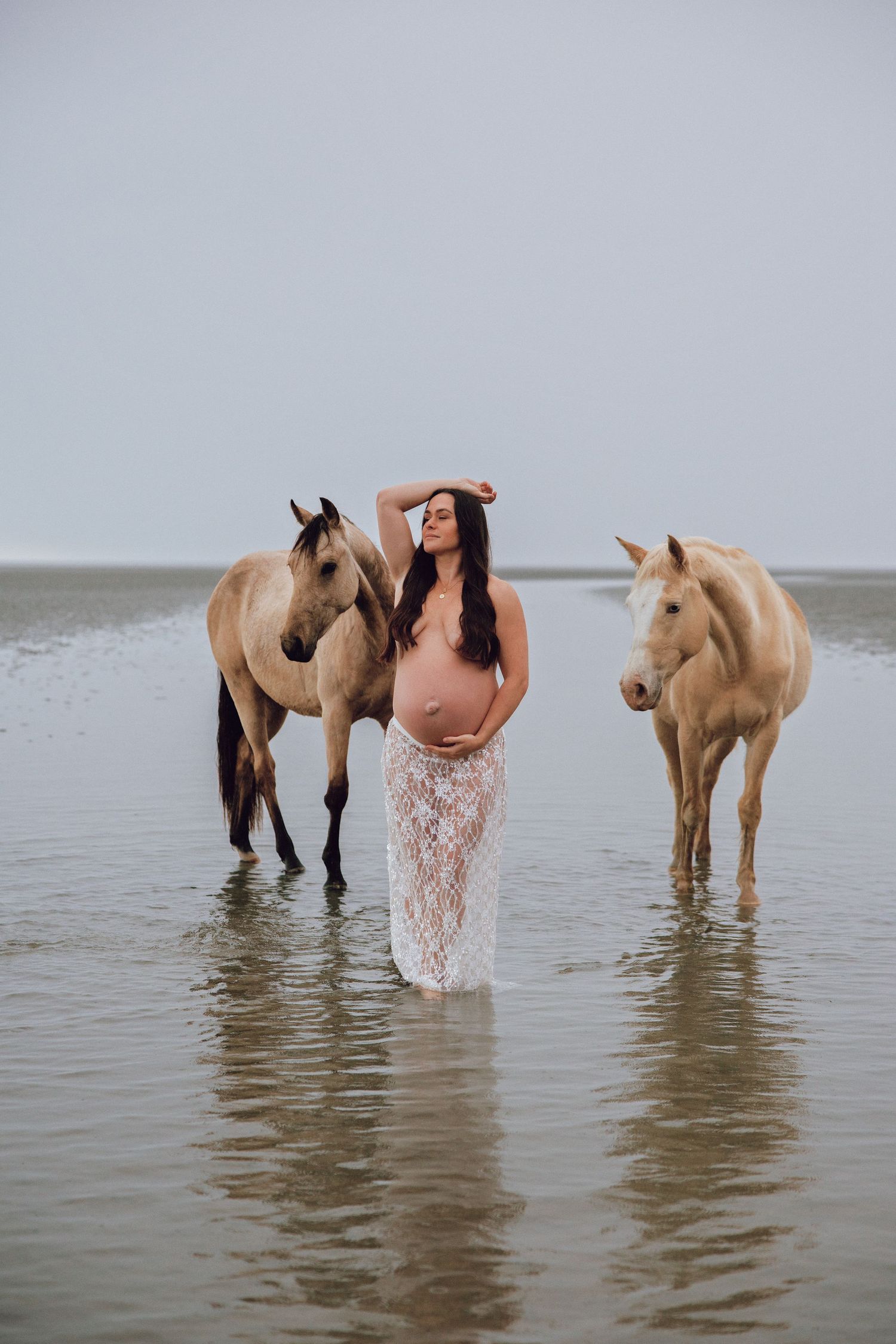 Pregnant woman in lace skirt poses between two horses in shallow water on a misty beach.