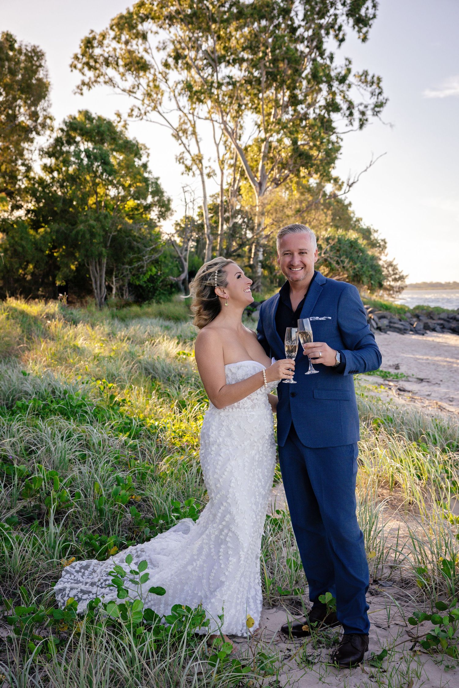 A couple toasts with champagne glasses at sunset on a beach with eucalyptus trees in the background.