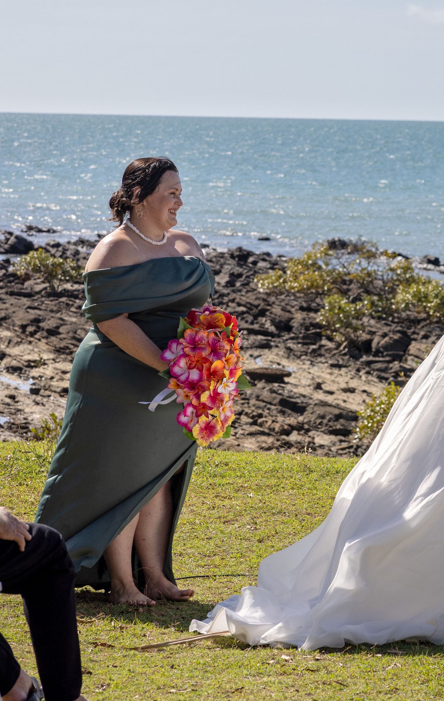 A wedding attendant in a dark green off-shoulder dress holds a vibrant pink bouquet on a scenic oceanfront cliff.