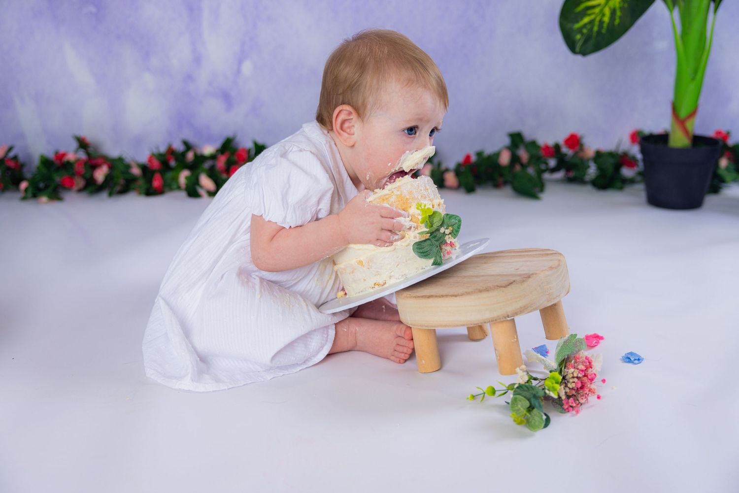 Baby in white outfit plays with colorful toys while lying on gray floor mat with floral backdrop.