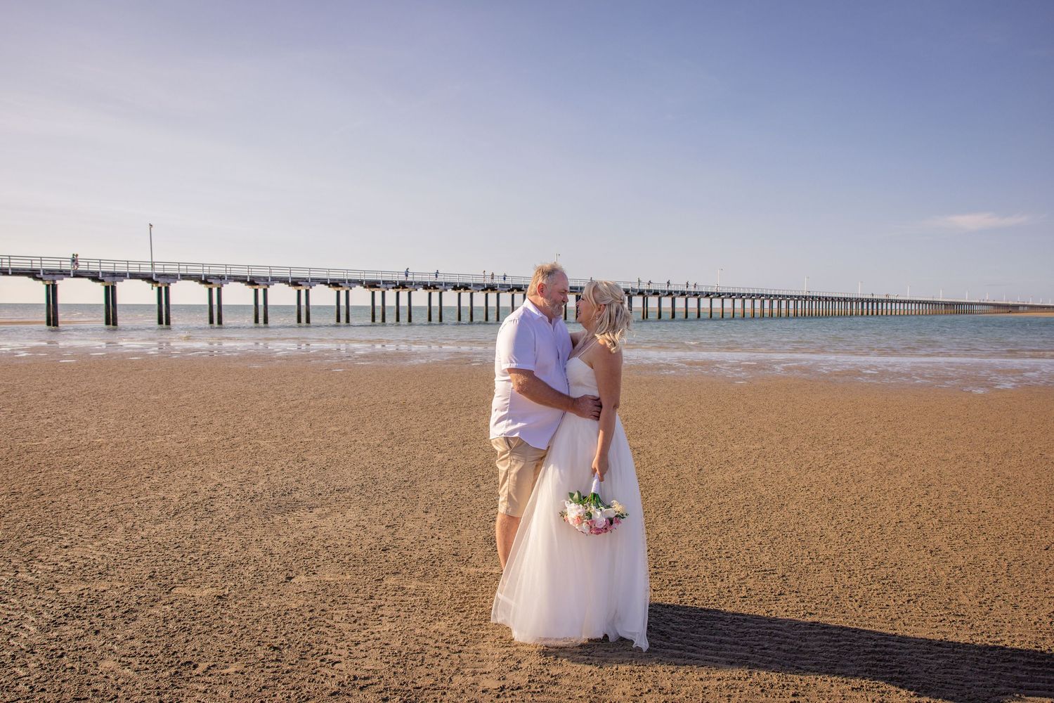 Wedding group poses on beach with long wooden pier stretching into the distance.
