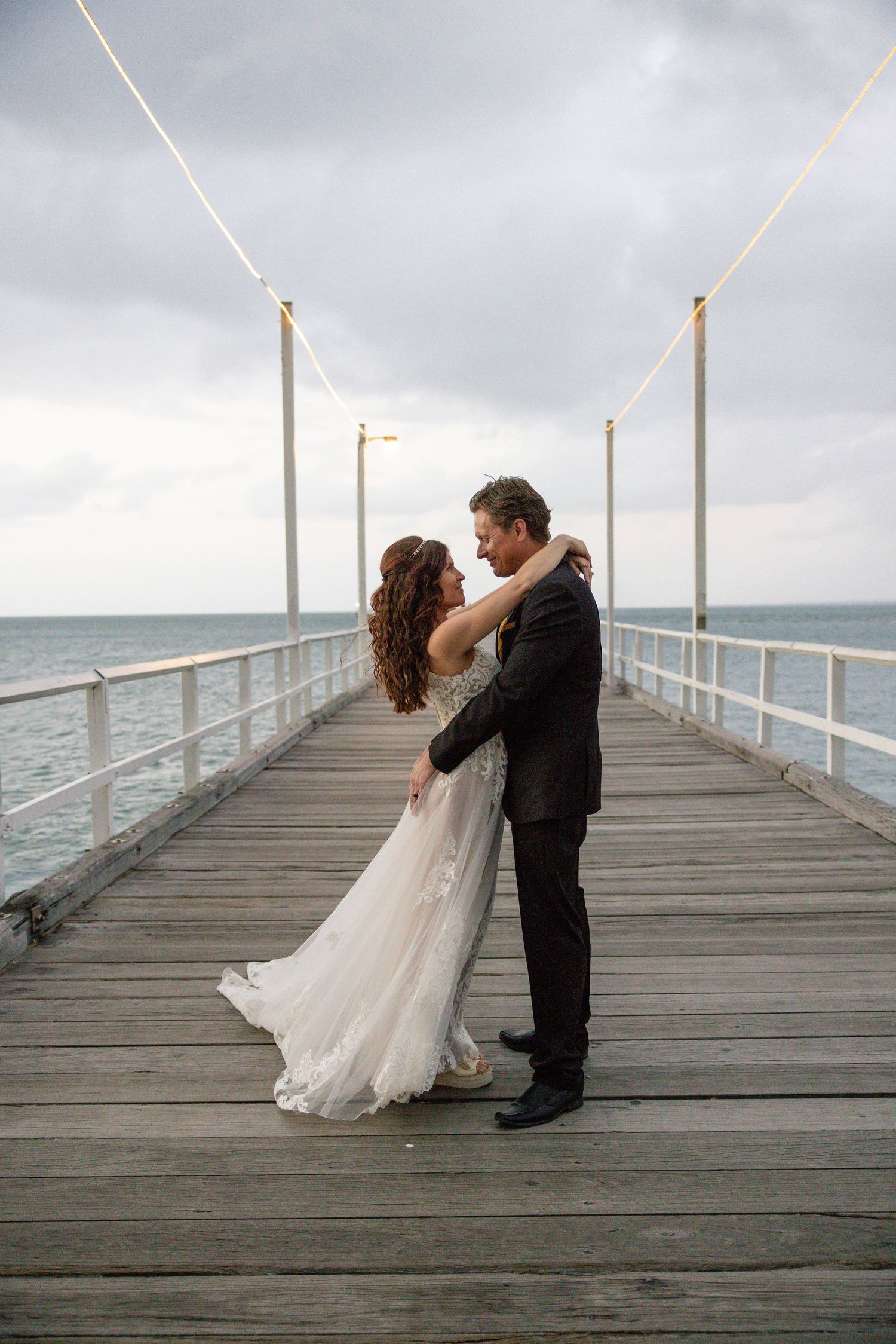 A romantic bride and groom share intimate dance poses on a wooden pier overlooking a stormy ocean.