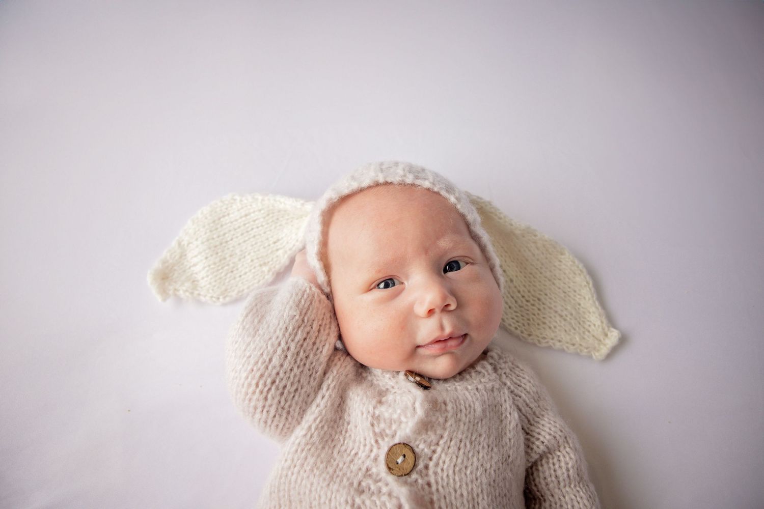 Baby wearing knitted bunny ear bonnet and cream sweater against light background.