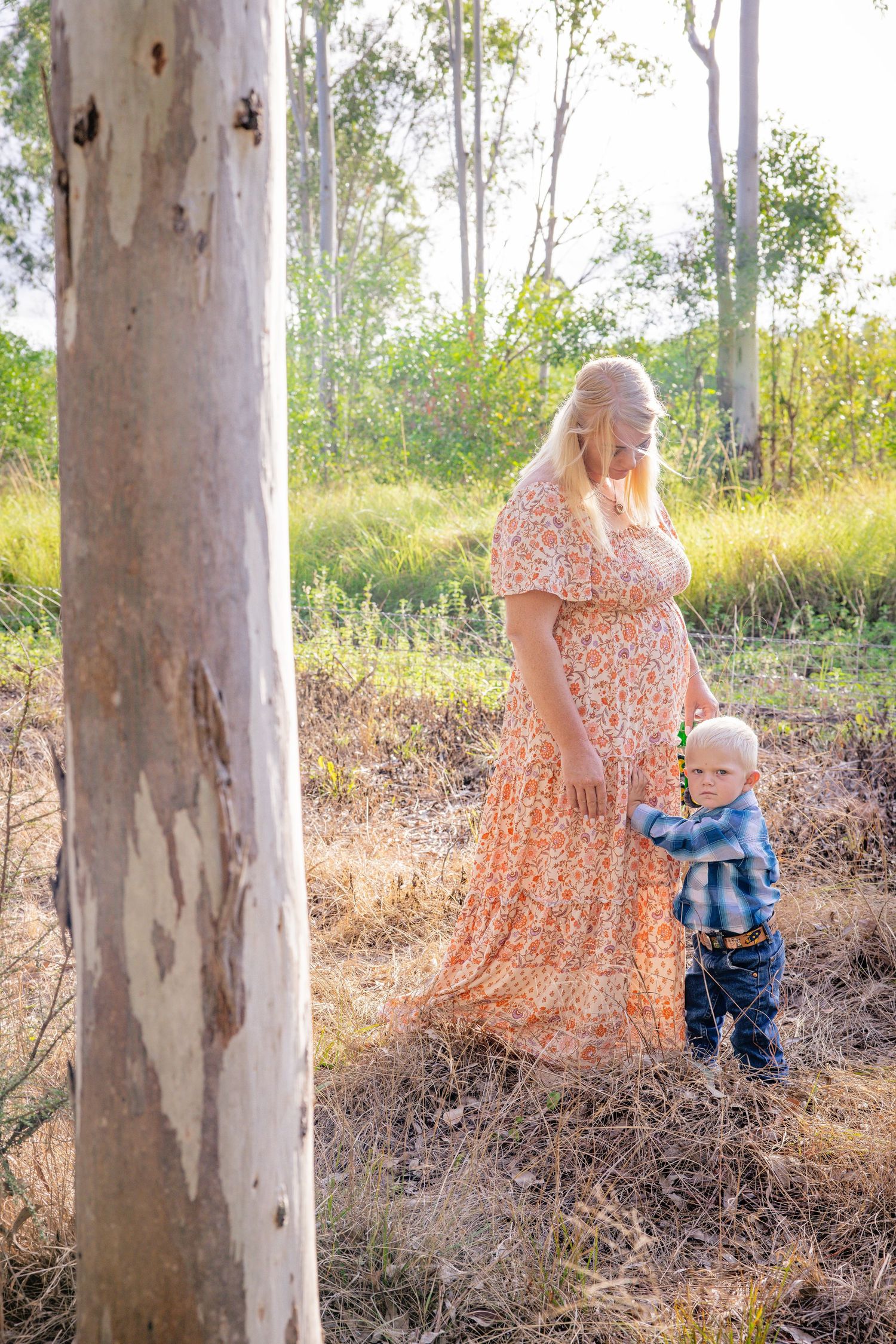 Expecting mother in floral dress stands by eucalyptus tree with toddler in outdoor setting.