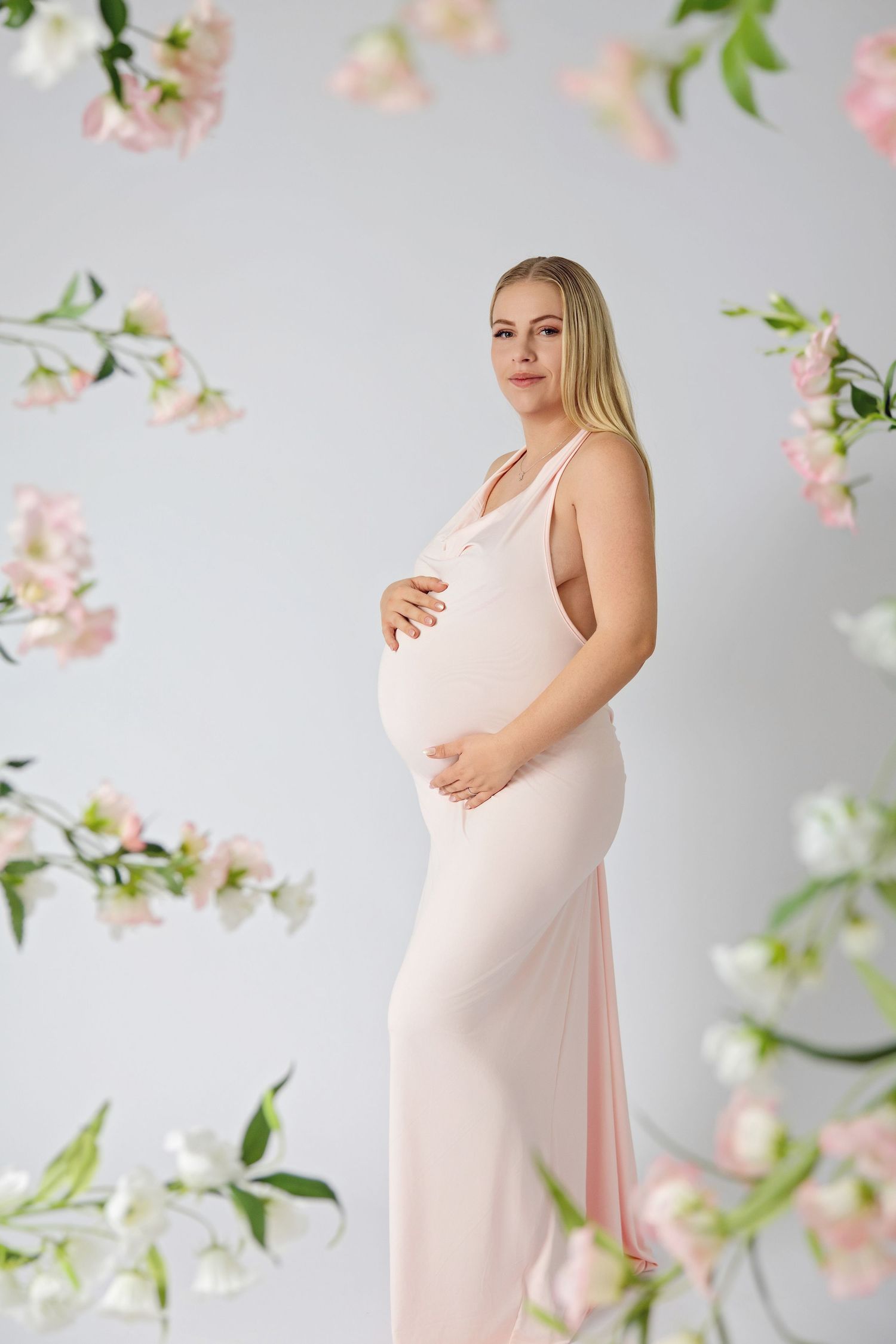 Pregnant woman in pink dress cradling baby bump surrounded by pink and white flowers on light background.