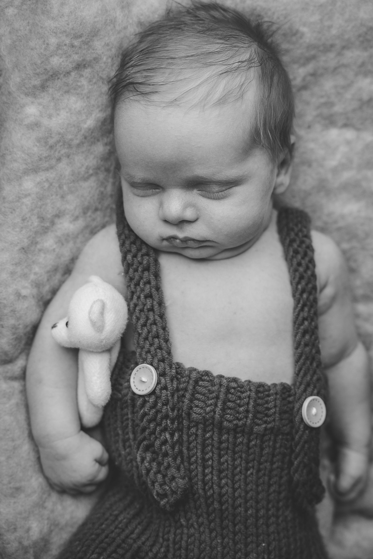 Black and white photo of a sleeping newborn baby in knit overalls holding a small white teddy bear.