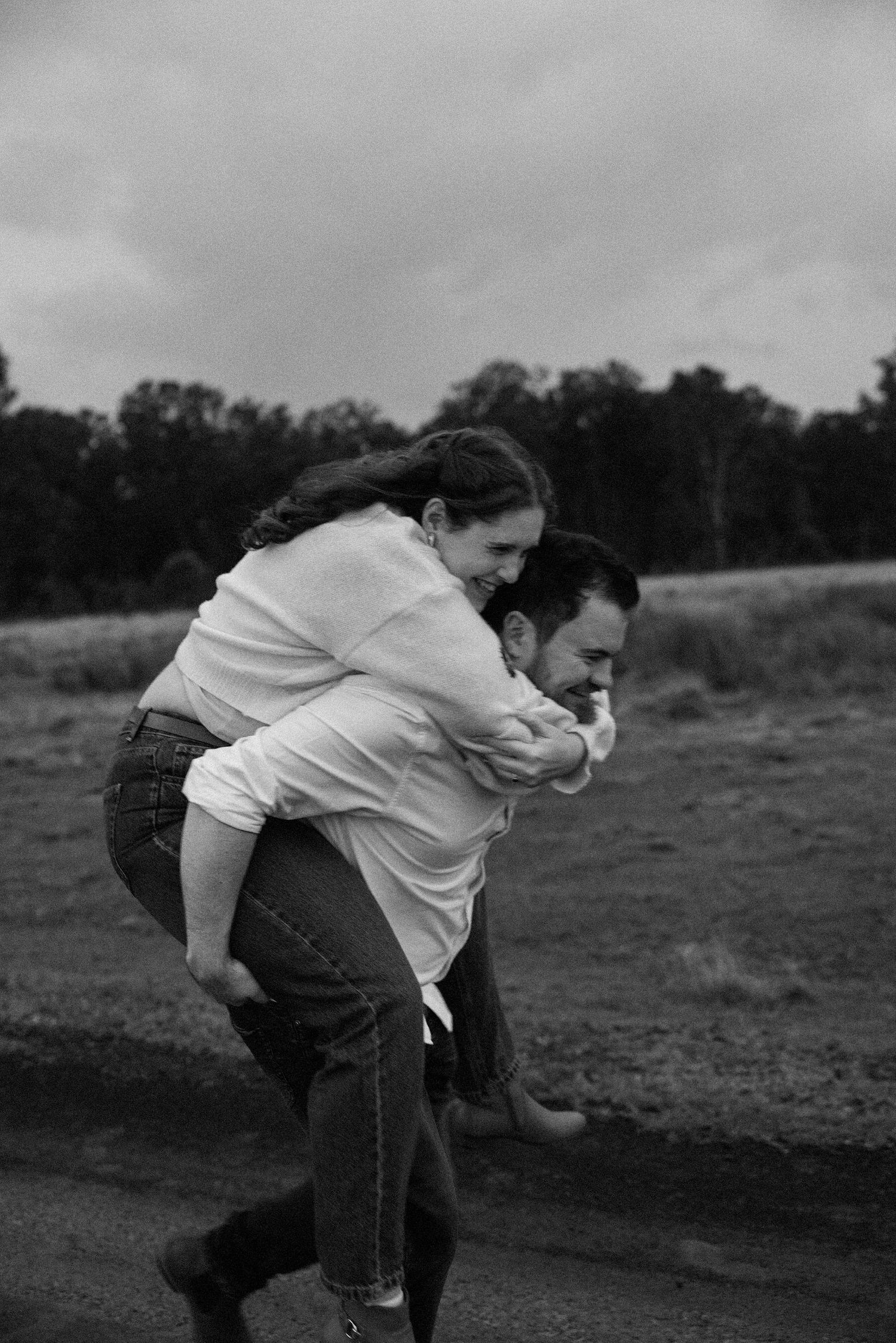 Black and white photo of a man giving a woman a piggyback ride outdoors, both laughing joyfully.