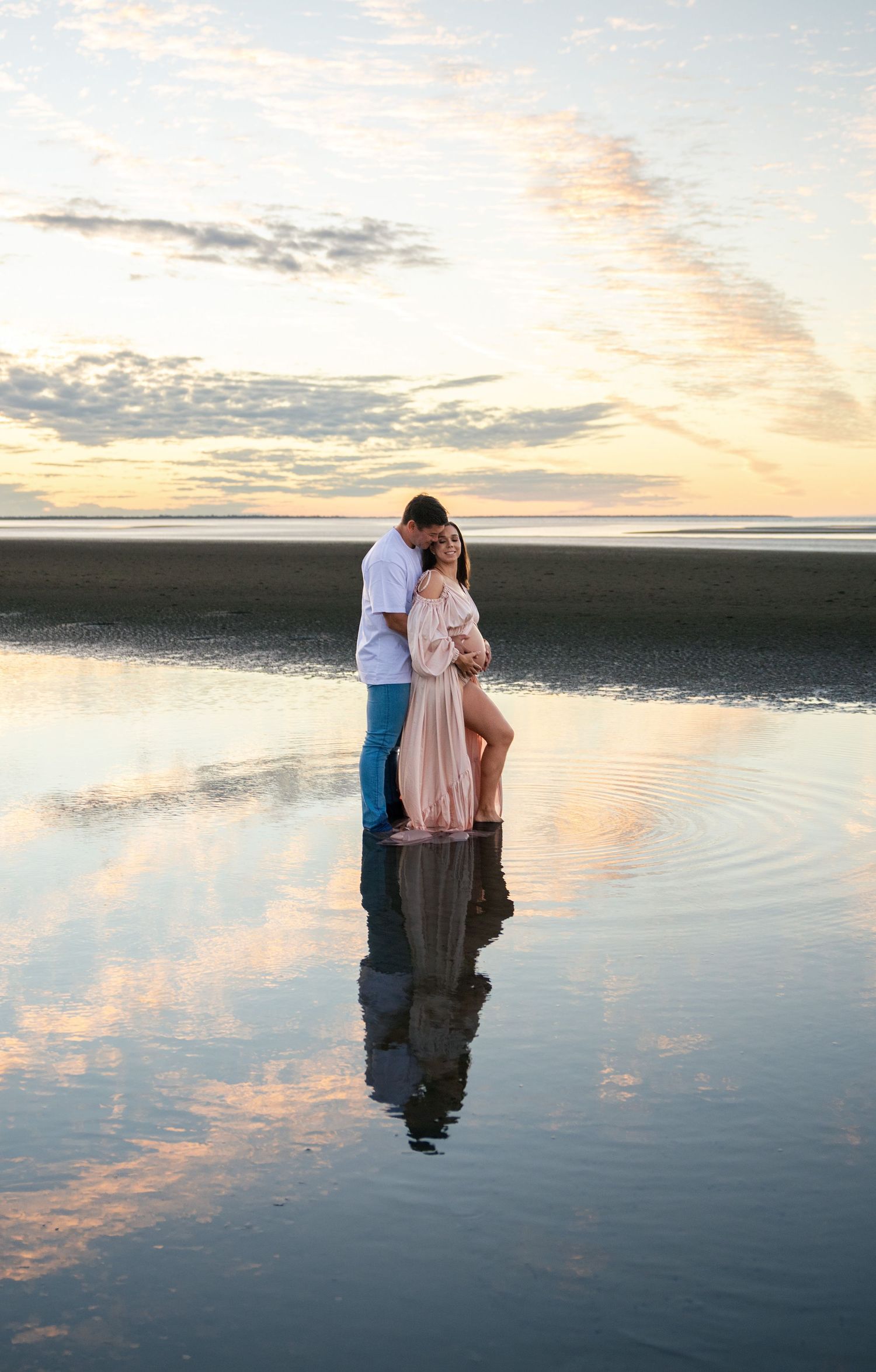 Couple embracing at sunset on a reflective beach, woman in pink maternity gown, romantic coastal maternity photo session.