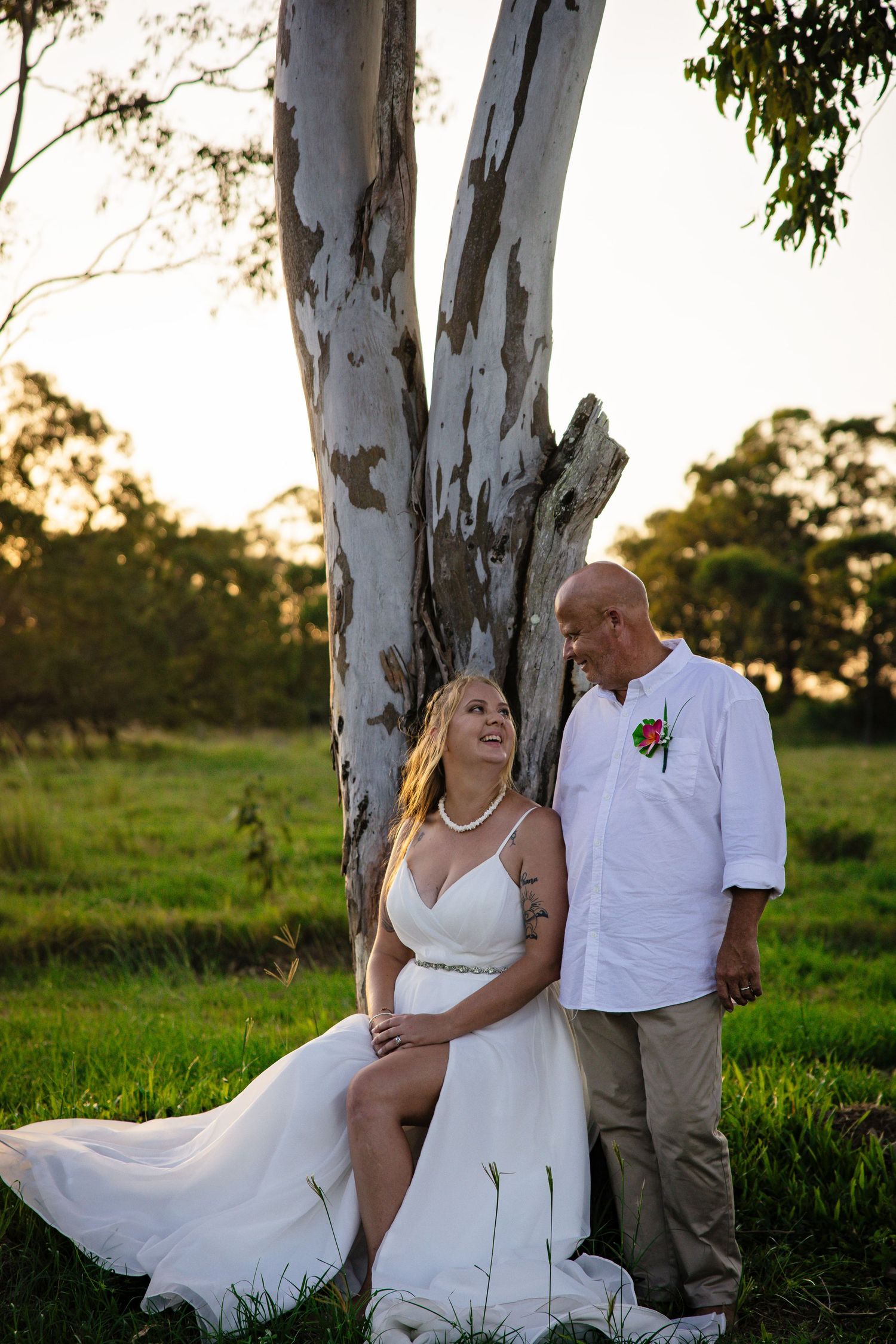 A romantic wedding photo at sunset features a couple posing together by a eucalyptus tree in a grassy field.