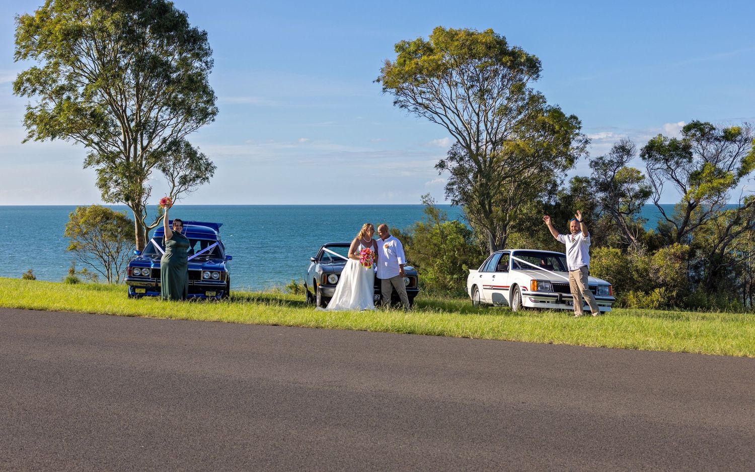 People gather on a coastal road overlooking the ocean with parked cars and tall eucalyptus trees framing the scenic view.