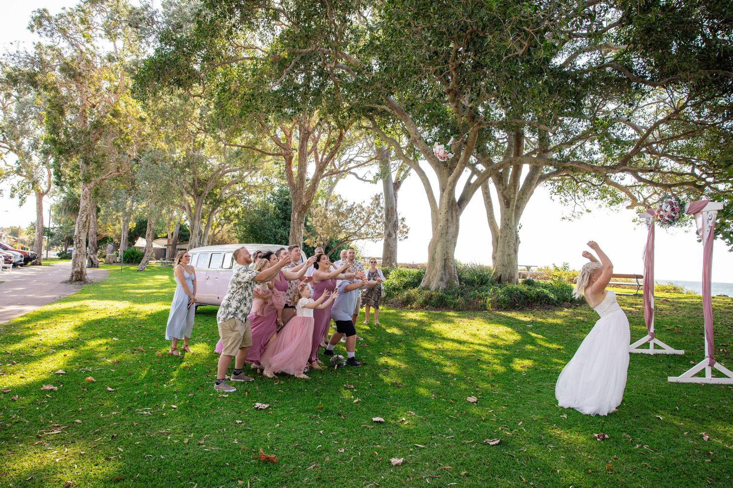 Wedding party gathers under shady trees at an outdoor ceremony venue during golden hour.