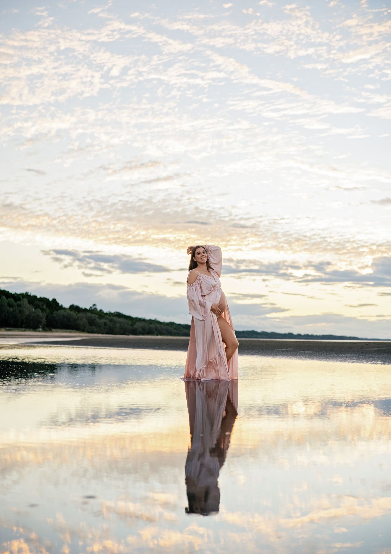 Woman in pink dress standing on reflective water at sunset with cloudy sky and tree line in background.