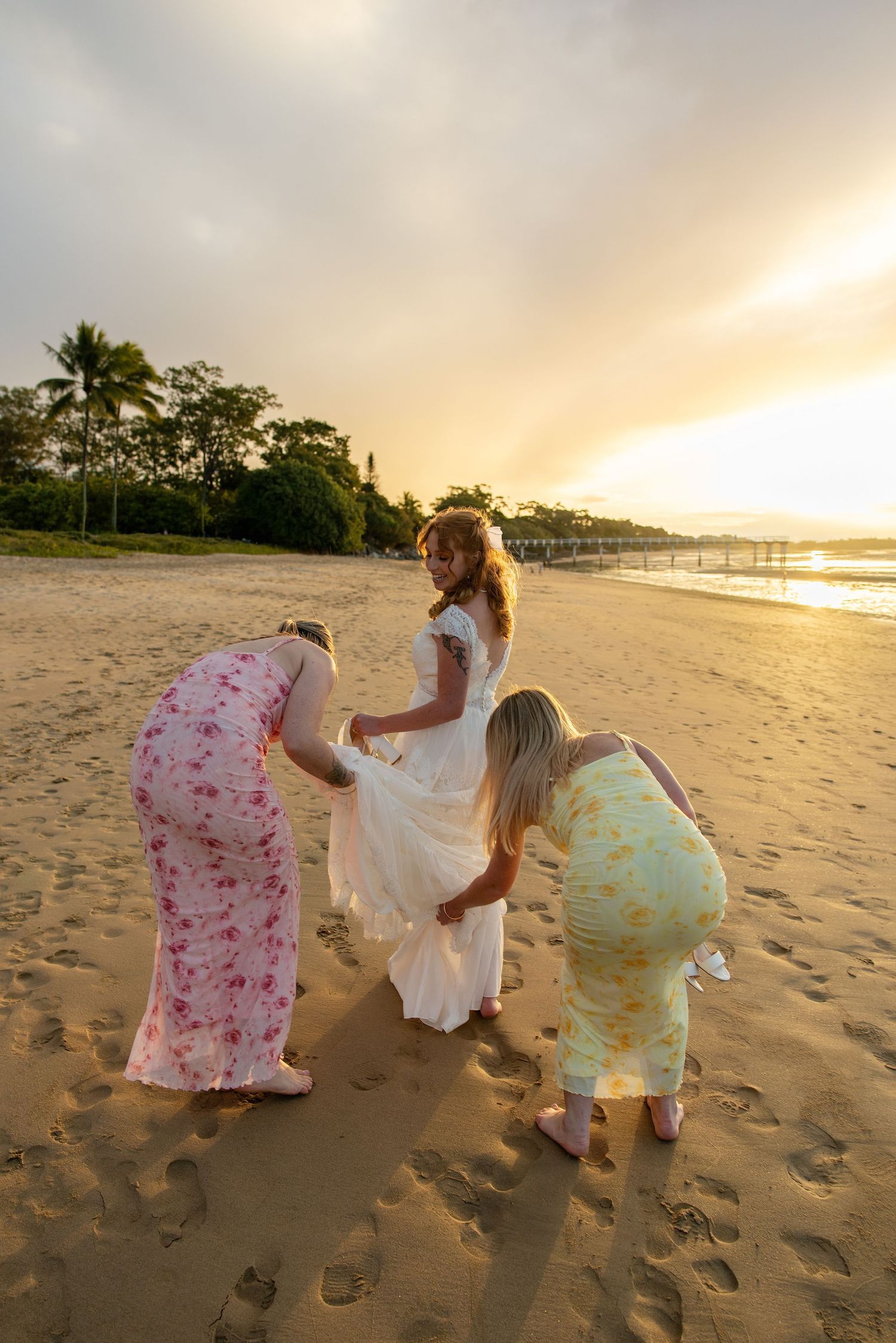 Bride in white gown assisted by two women in floral dresses on sandy beach at golden sunset.