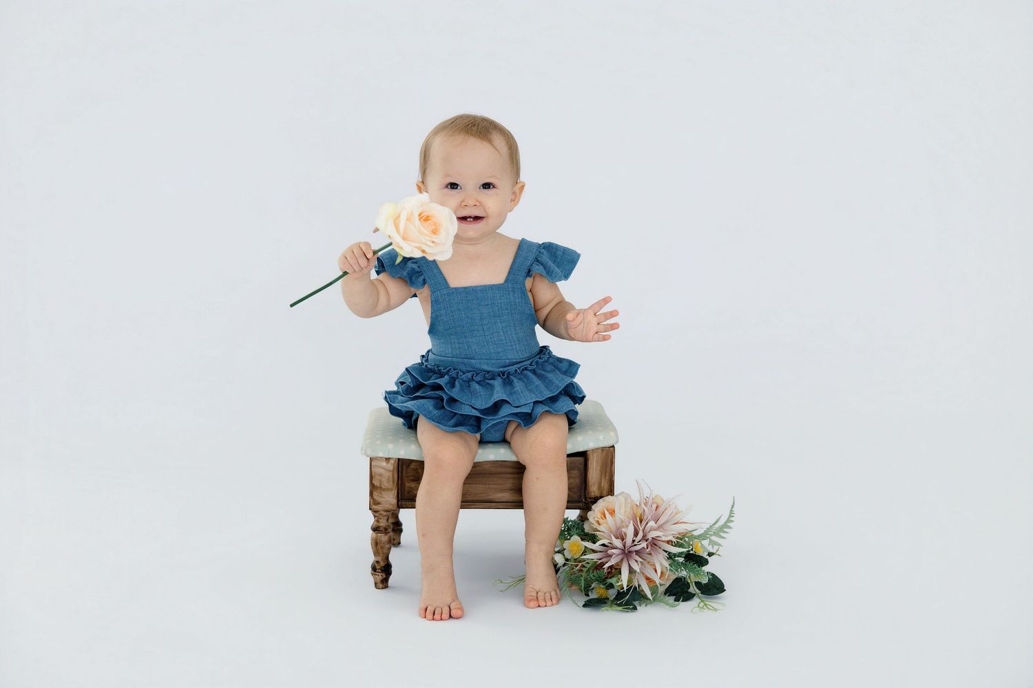 A smiling baby girl in a blue ruffled romper sits on a small chair holding a flower, with blooms on the floor beside her.