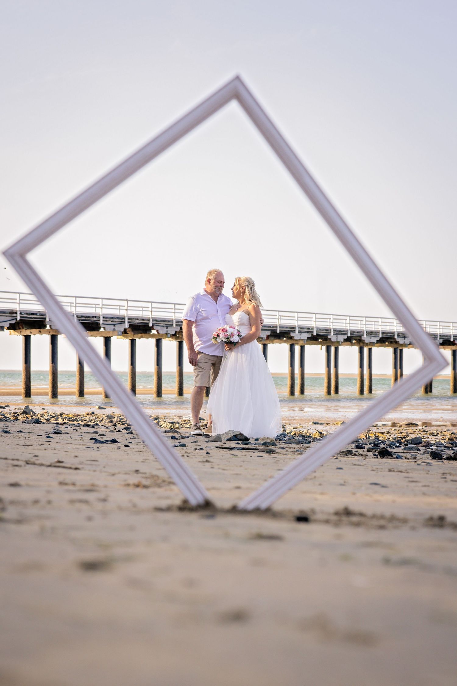 A series of creative beach wedding photos taken through white geometric frames with a pier in the background.