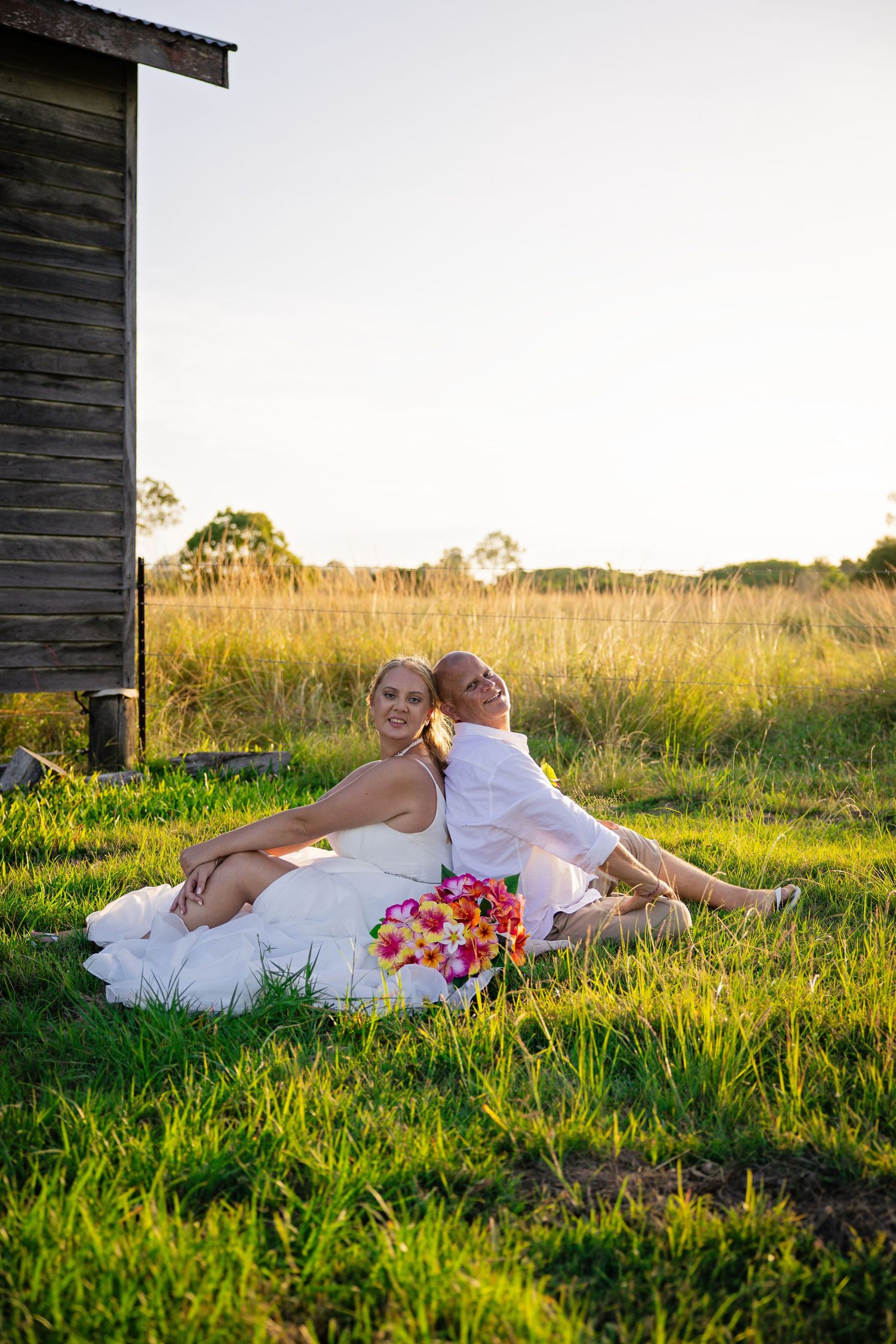 Couple in white outfits relaxing together on green grass near rustic barn at golden hour.