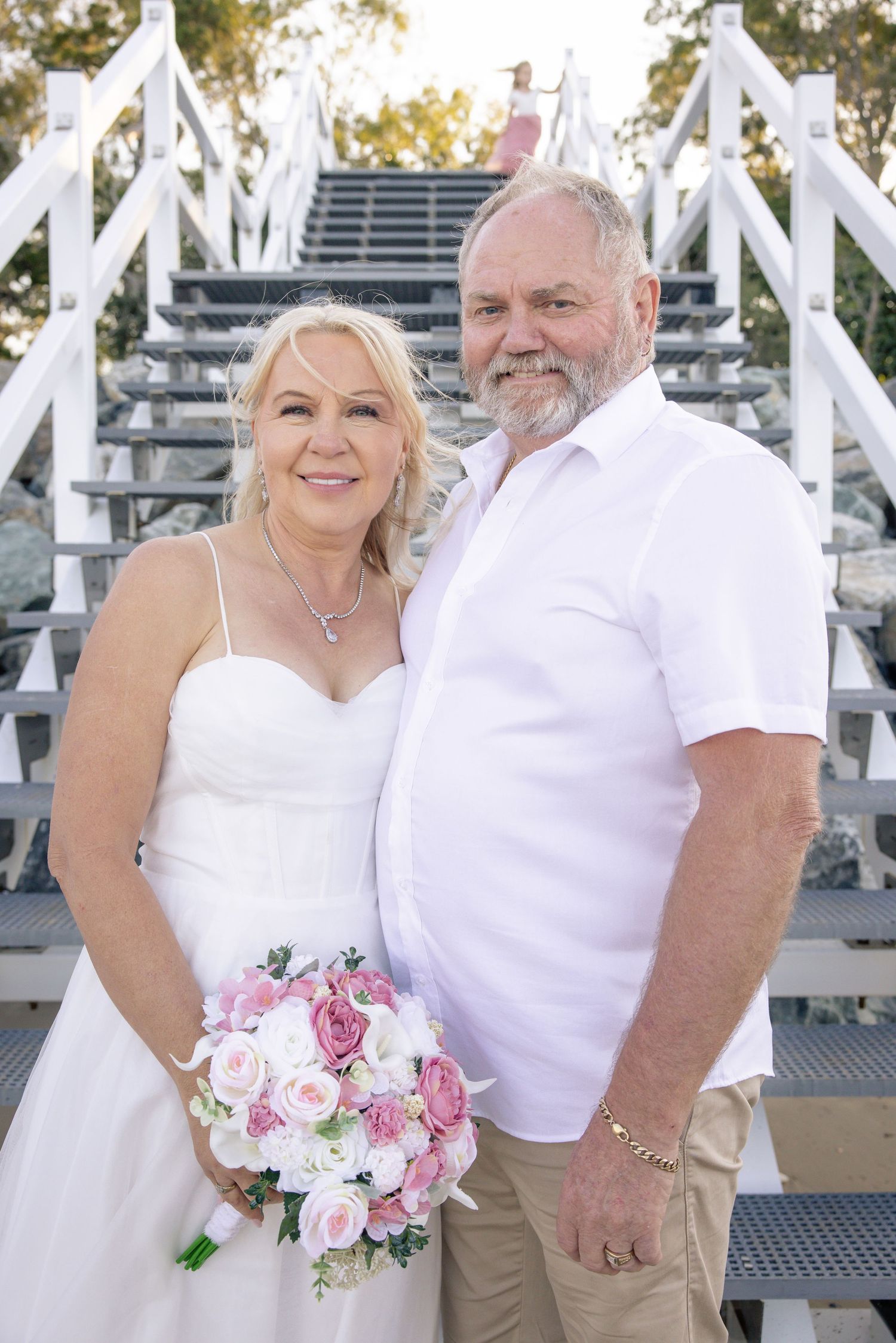 A couple in white wedding attire pose together on a white lattice deck with pink and white floral bouquet.
