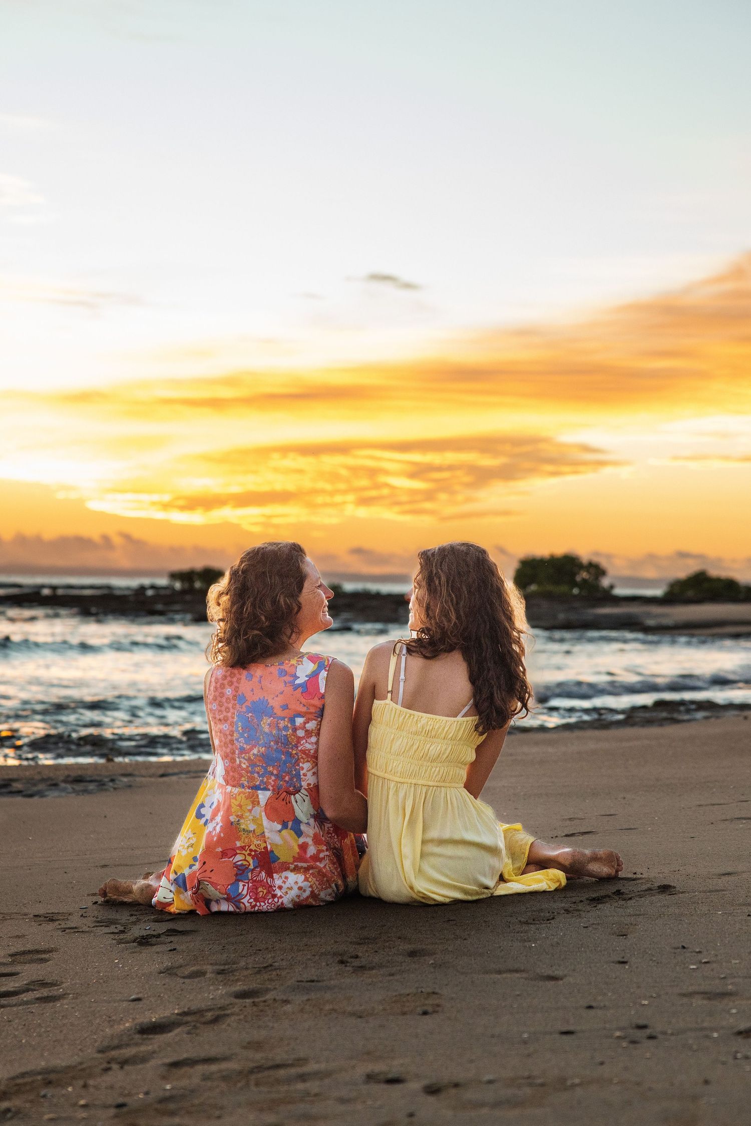 Two women in colorful dresses sit on a sandy beach watching a vibrant golden sunset over the ocean.