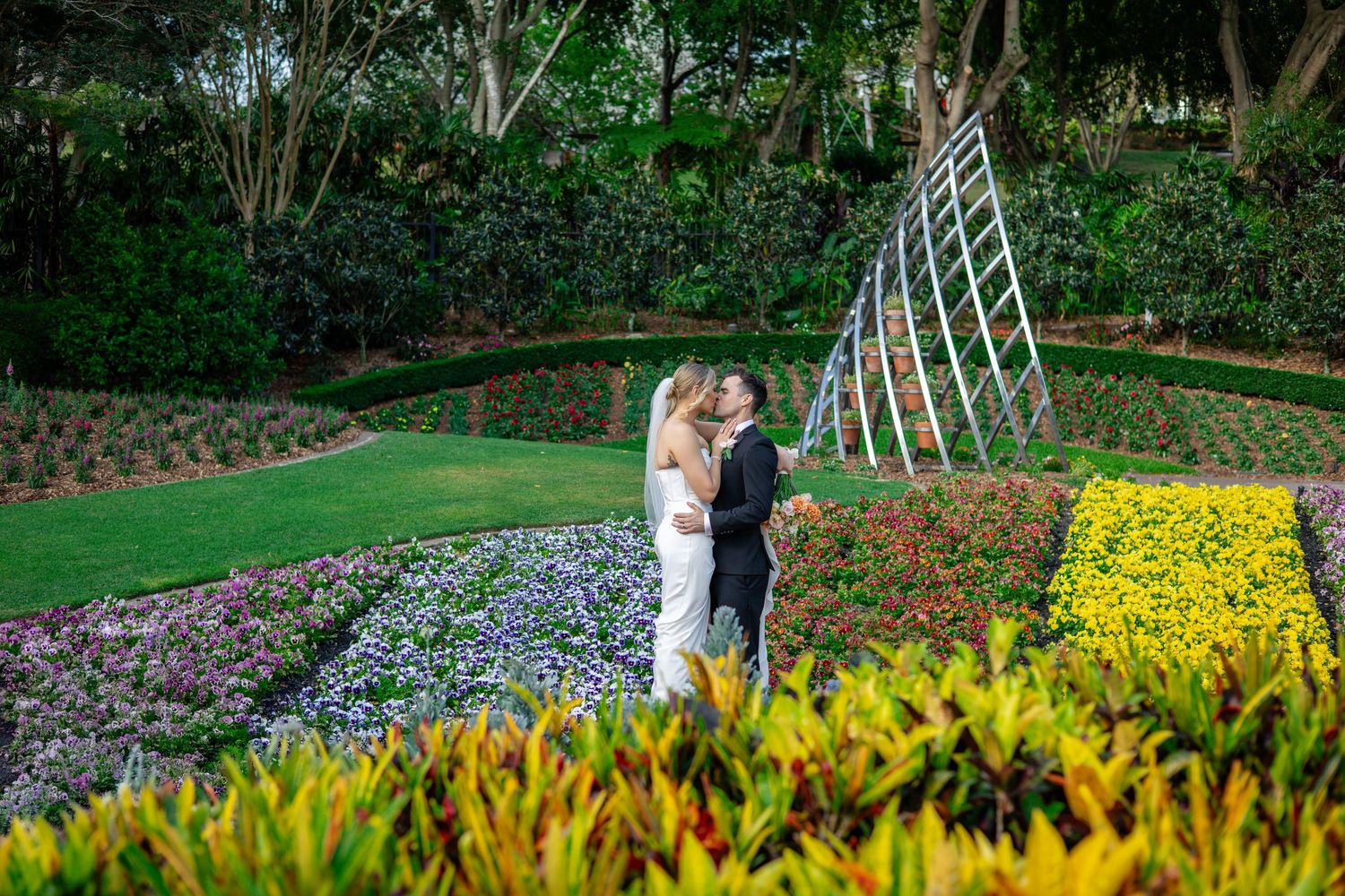 A couple embraces in a vibrant garden with colorful flower beds and a decorative lattice structure in the background.