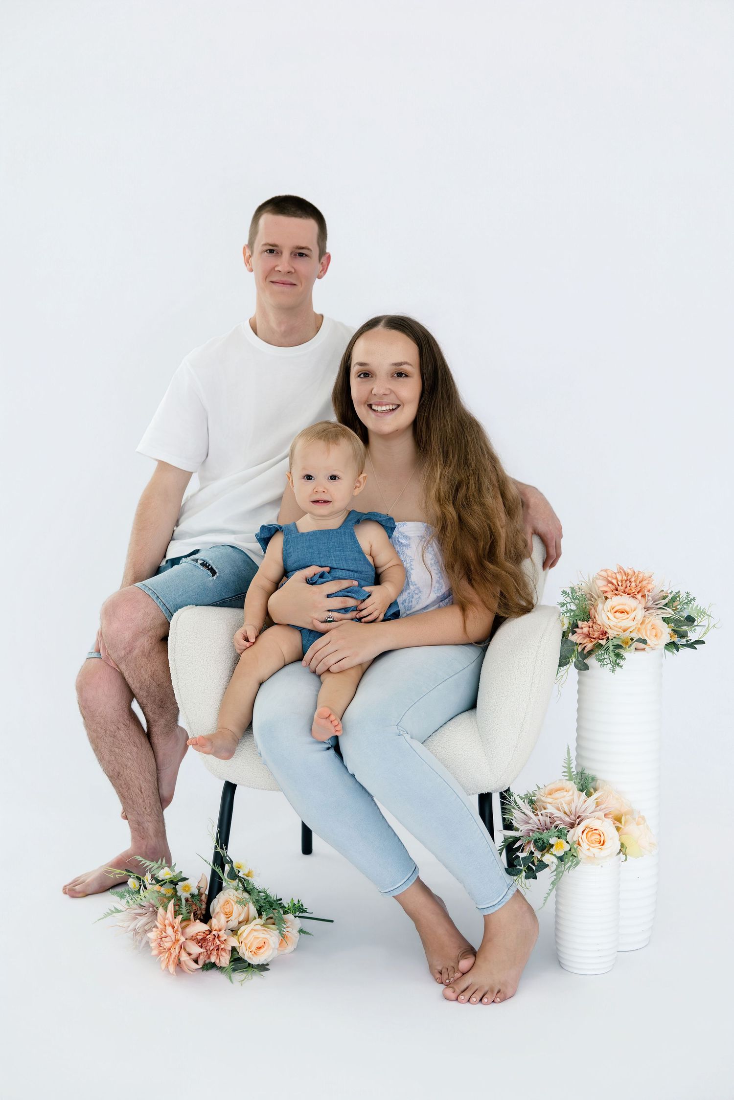 Young family with baby in blue outfit posing on white chair with flowers in bright studio setting.