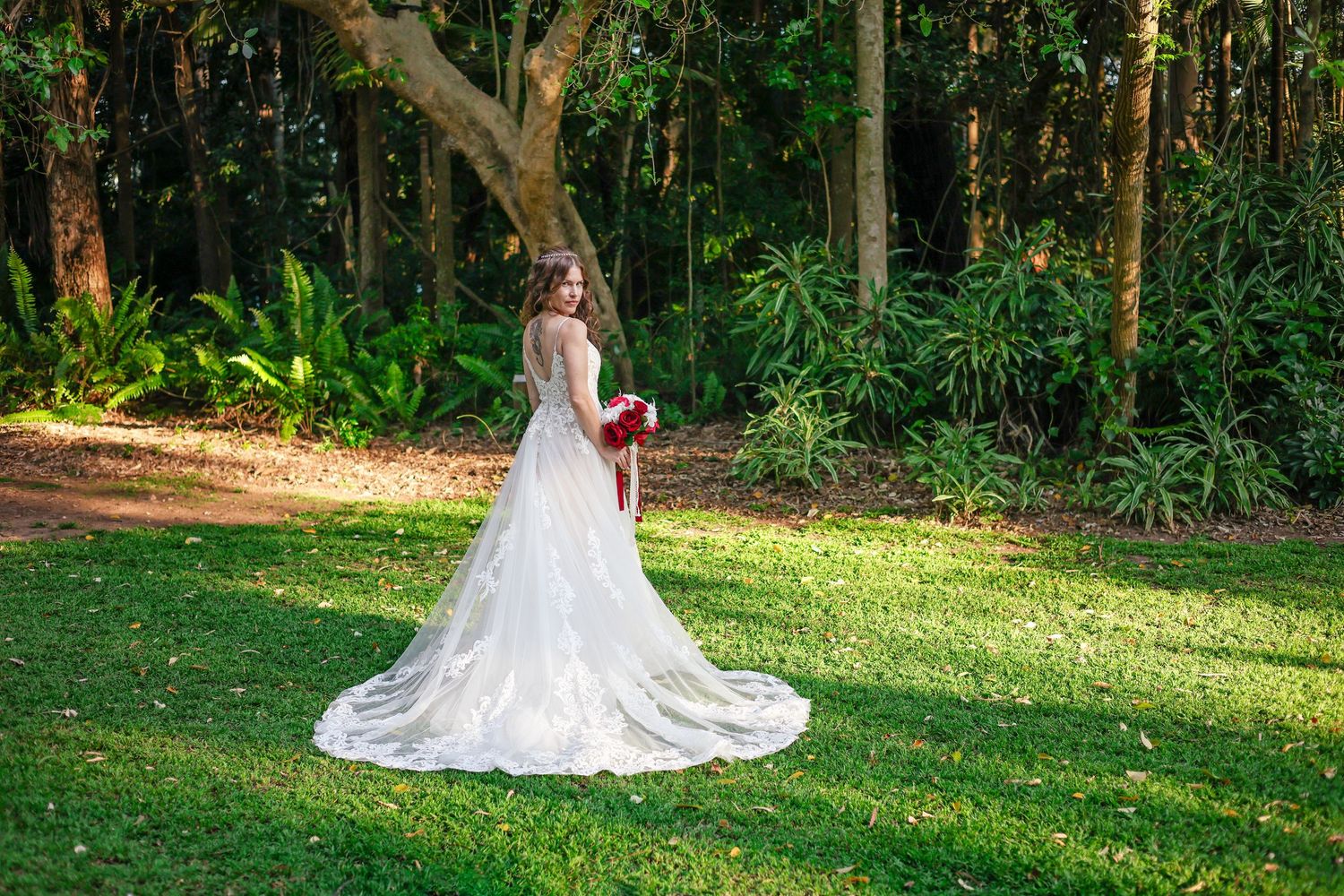 Bride in elegant white gown with long train holding red bouquet, standing on green lawn surrounded by lush forest.