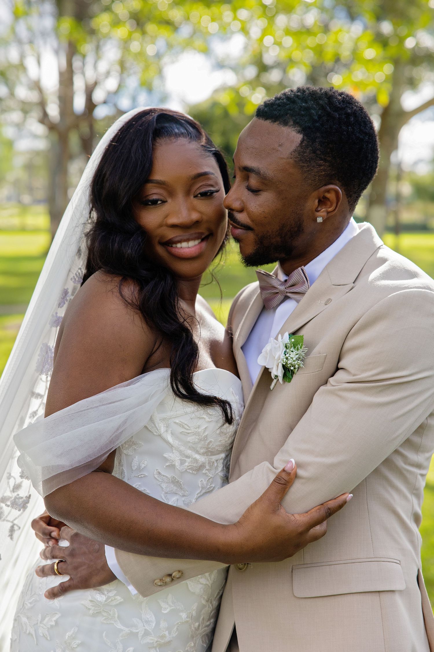 A joyful bride in a white gown and veil embraces her groom in a tan suit with bow tie outdoors.