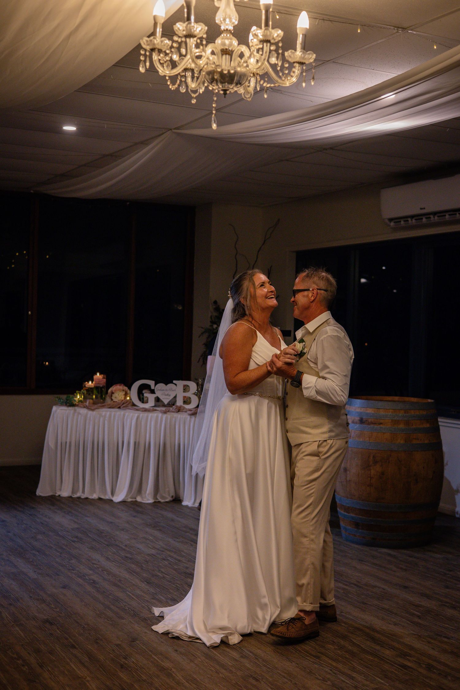 A couple shares an intimate dance moment under elegant chandeliers in a rustic reception venue with wooden floors and barrels.
