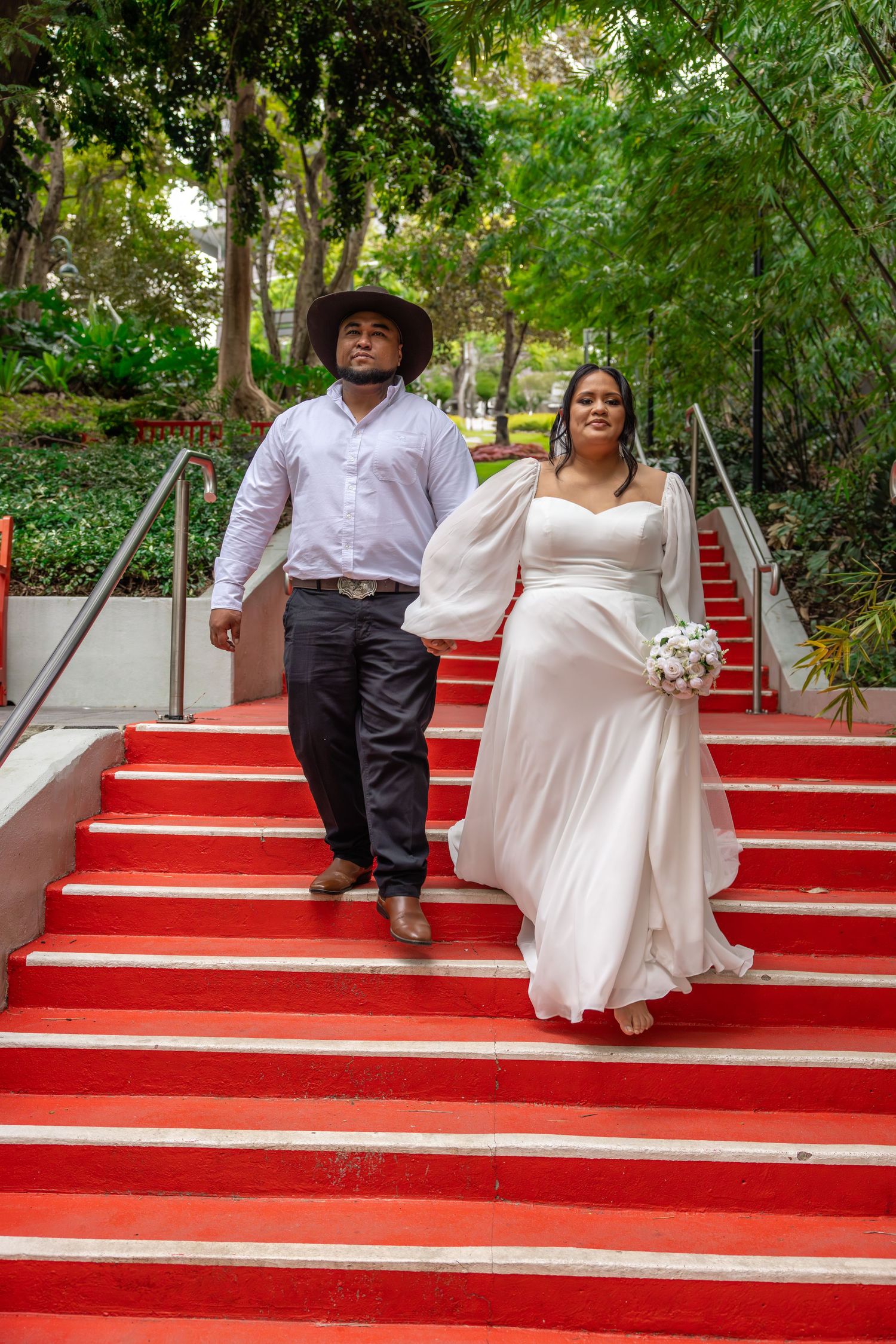 A couple descends bright red outdoor stairs surrounded by lush greenery, with one wearing a wedding dress and holding a bouquet.