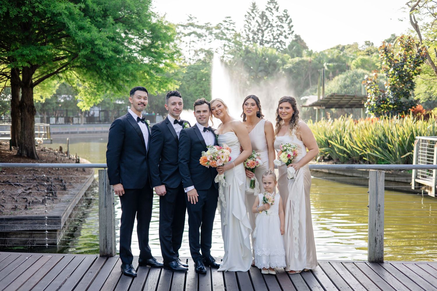 Wedding party poses together on wooden dock by scenic lake during outdoor celebration.