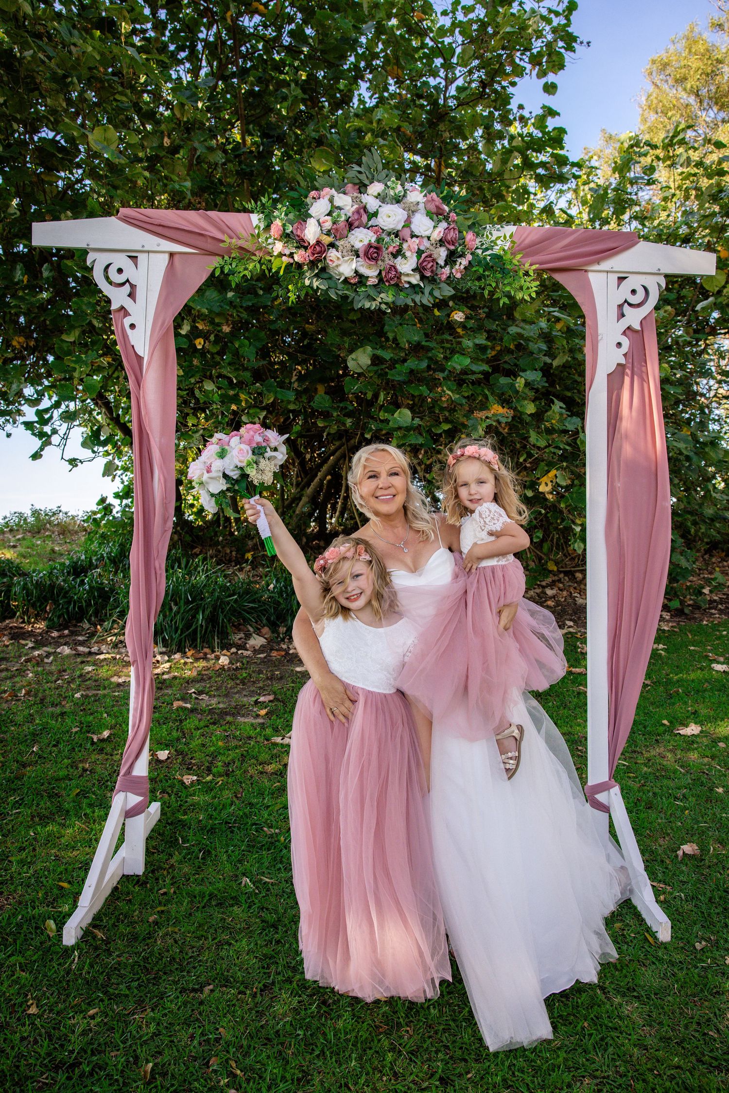 Outdoor wedding scene with pink and white dresses against a lush garden backdrop with decorative arches.