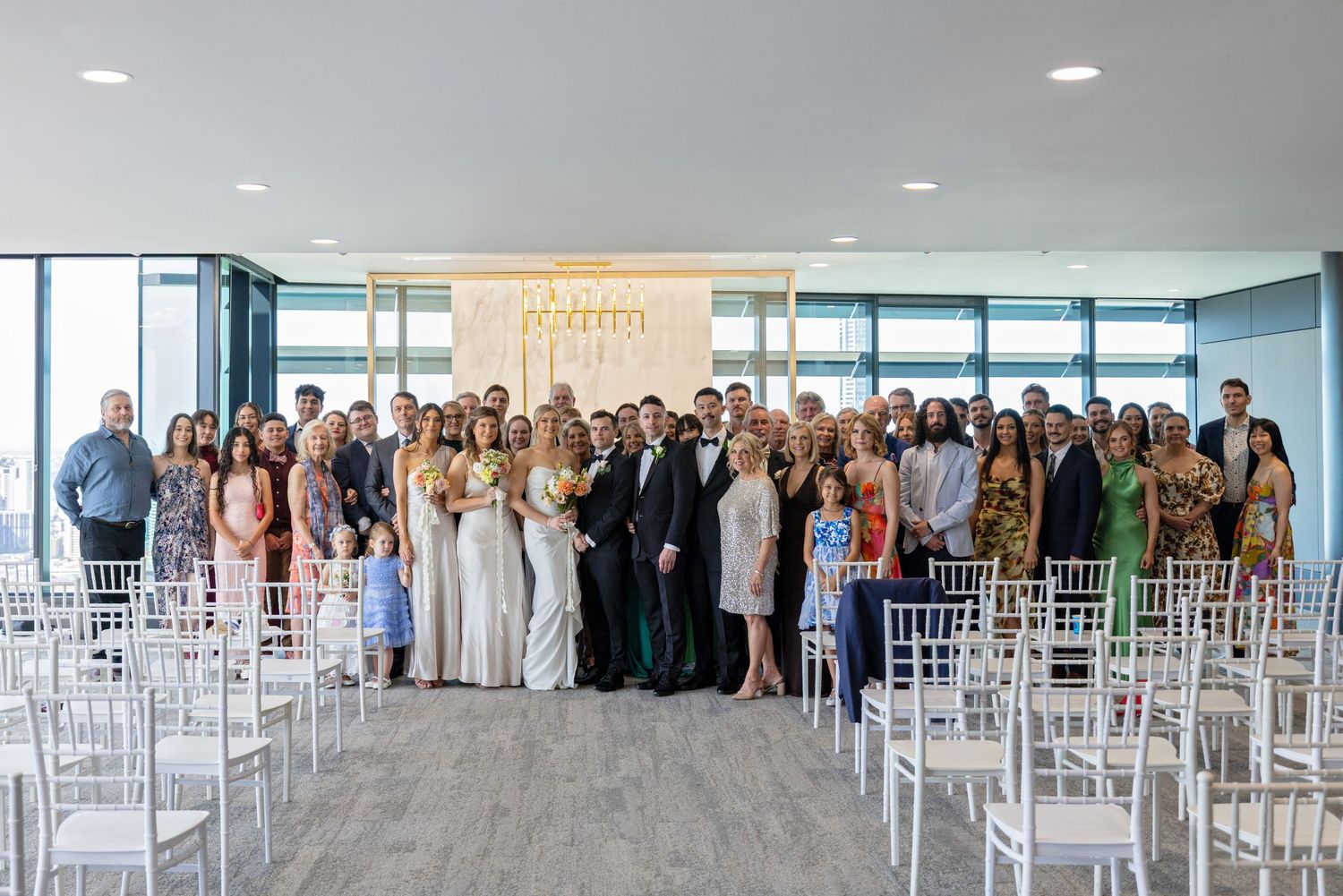 Large wedding party poses together in modern venue with white chairs and floor-to-ceiling windows.