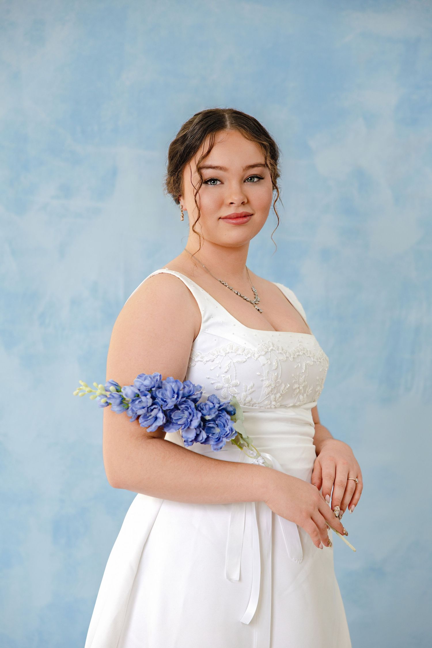Young woman in white bridal gown with blue floral corsage poses gracefully against a soft blue backdrop.