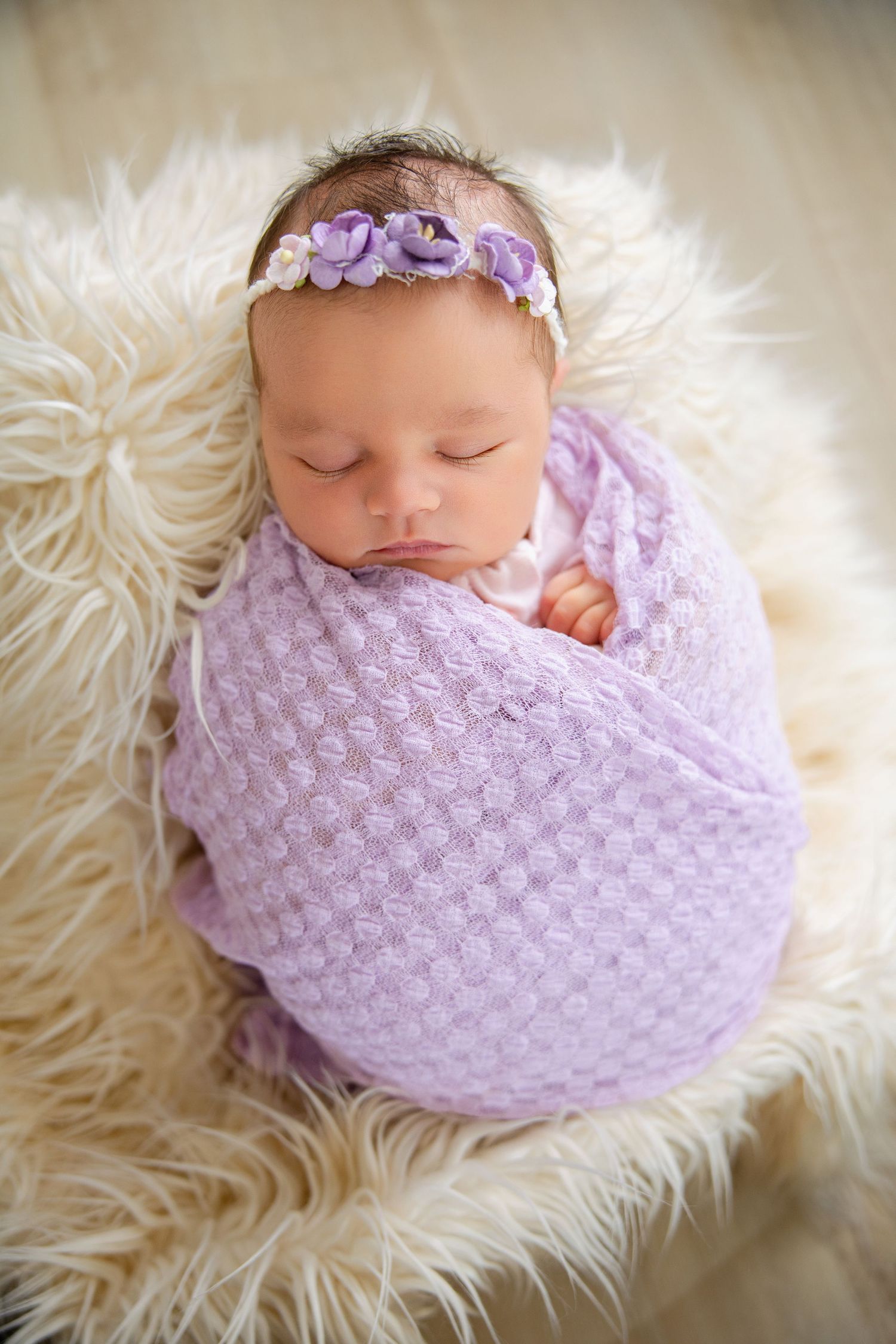 Sleeping newborn baby girl swaddled in purple knit wrap with floral headband on fluffy white blanket.