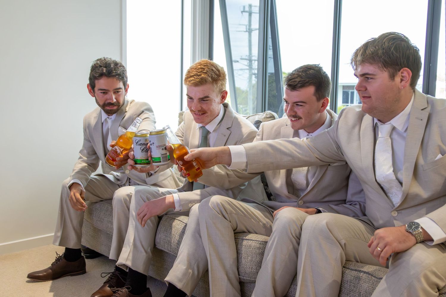 Groomsmen in light gray suits and orange boutonnieres sit together during relaxed wedding celebration photo session.